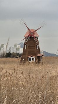 Rustic windmill stands in a dry field against a backdrop of city buildings.