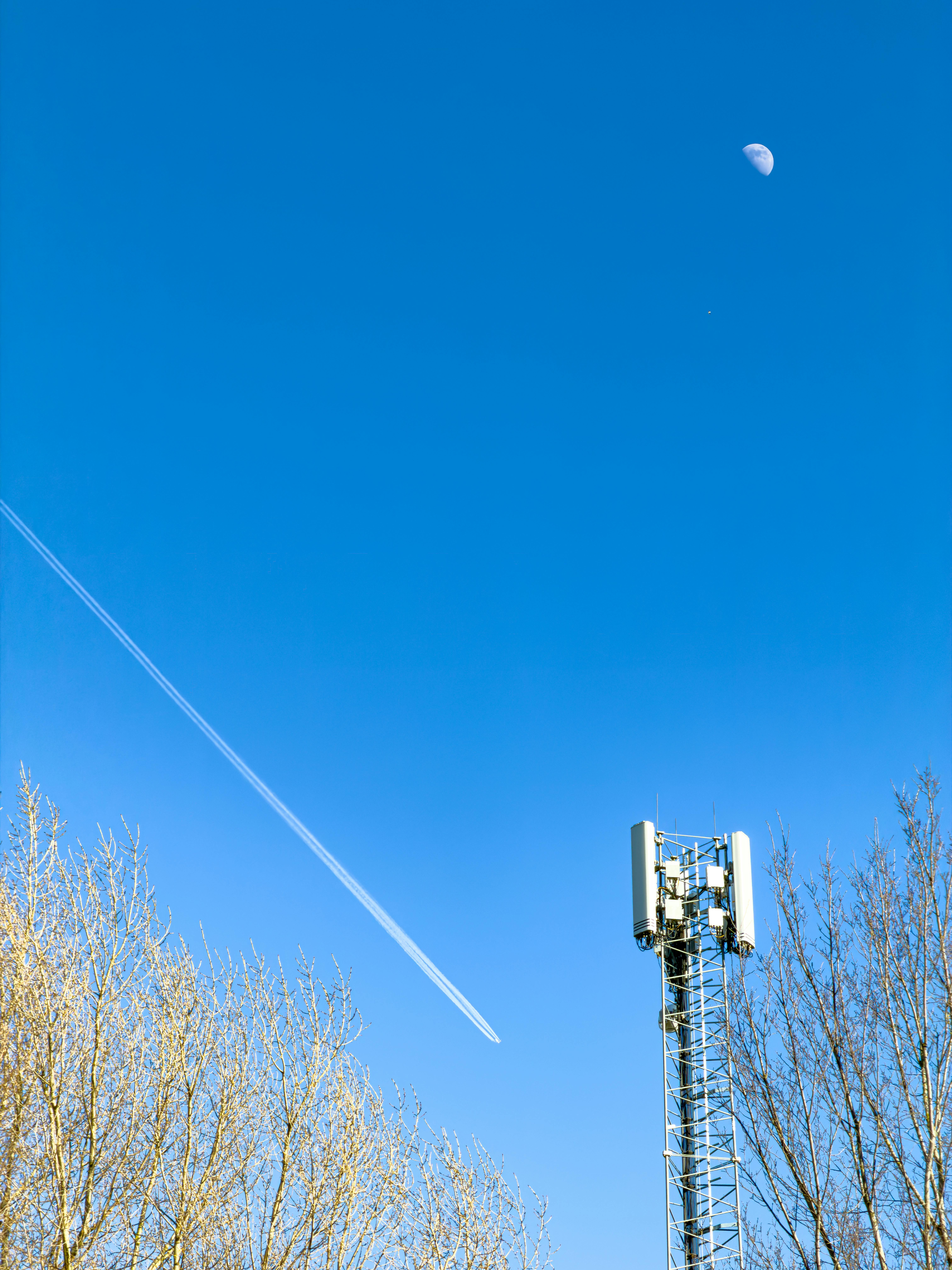 A cell tower reaching towards a clear blue sky with a visible contrail and moon.