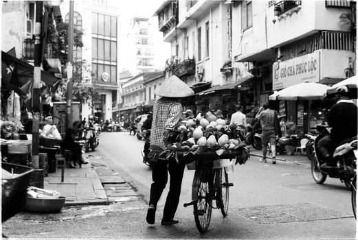Black and white photo of a street vendor pushing a bicycle with fruits in a lively urban scene.