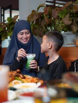 A mother in a hijab and her child enjoying a green drink and food indoors, surrounded by plants.