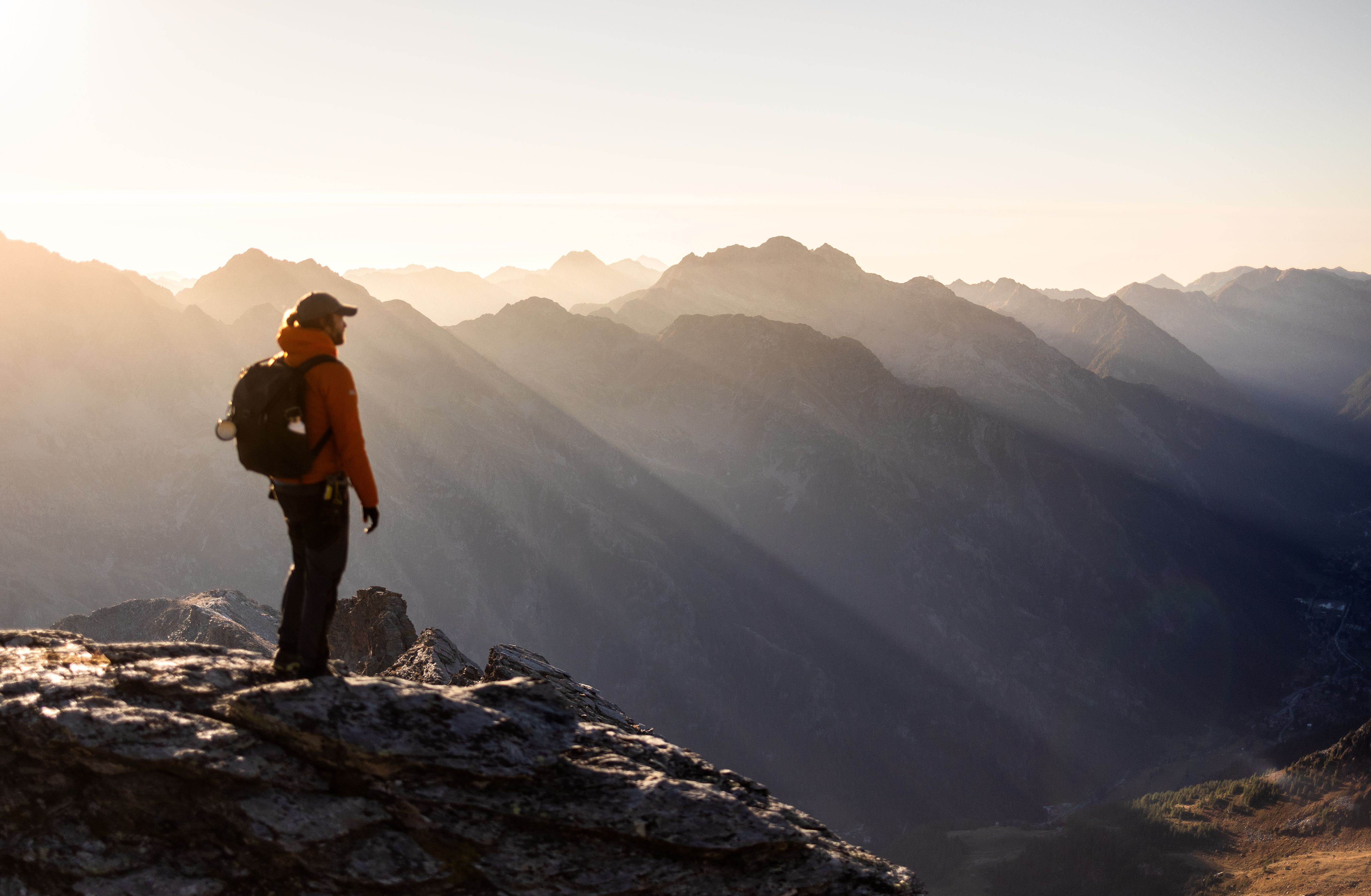 A solitary hiker in orange jacket stands on a rocky summit, admiring the sunrise over the mountain peaks.