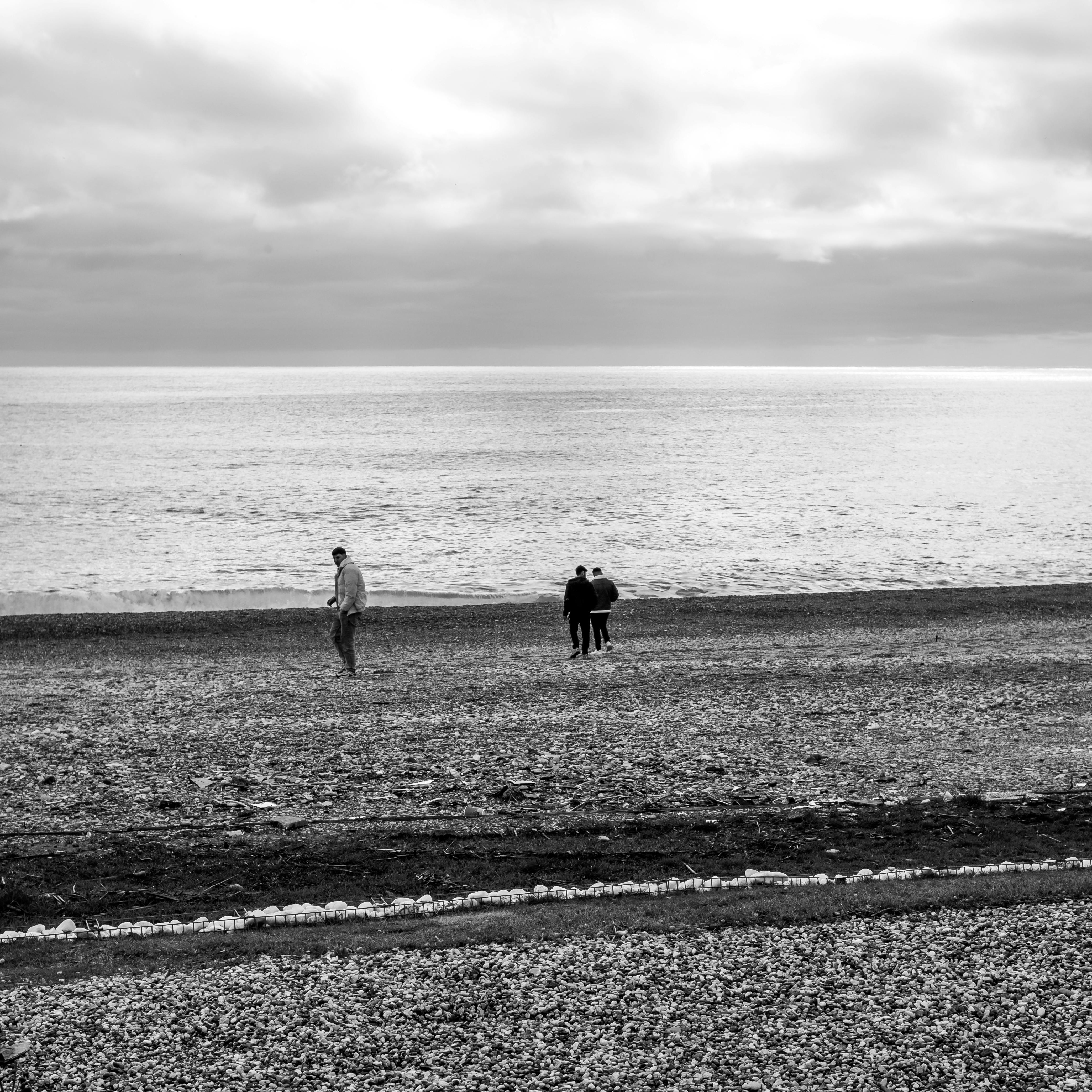gratis Zwart-witfoto van mensen die onder een bewolkte hemel over een kiezelstrand lopen. Stockfoto