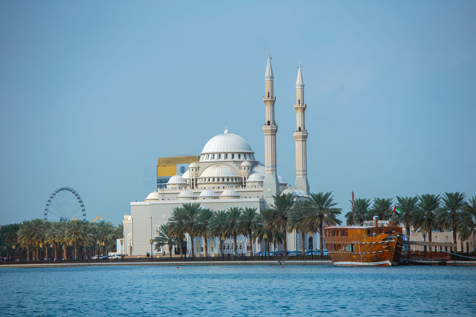 Scenic view of a mosque by the water with a dhow and palm trees in Sharjah, UAE.