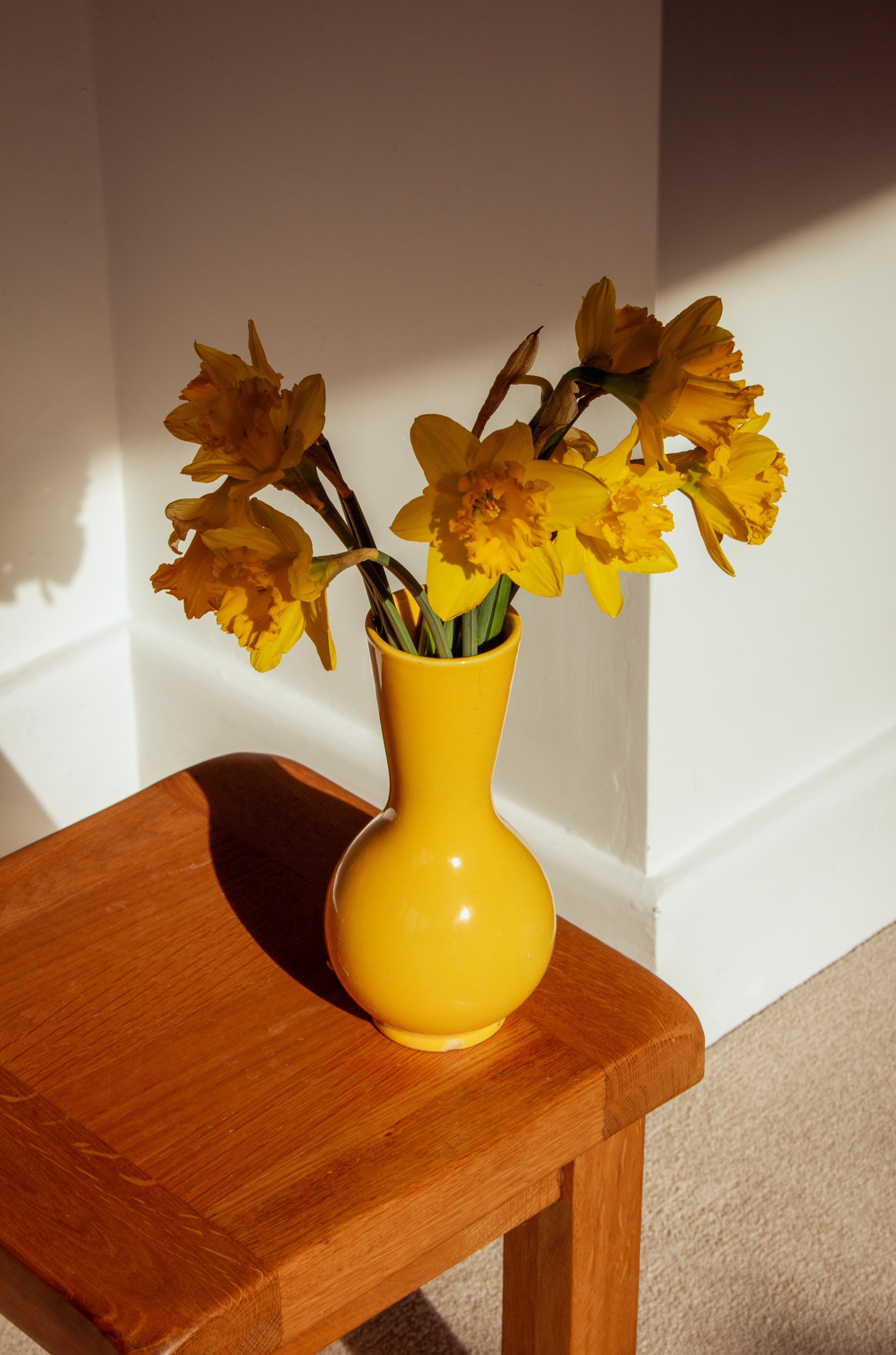 Bright daffodils in a yellow vase on a wooden table, illuminated by sunlight.