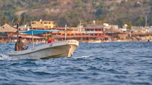 Free Two fishermen navigate a boat in vibrant coastal waters with a bustling village in the background. Stock Photo