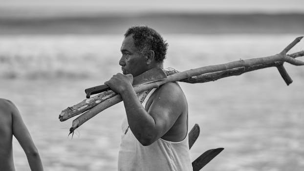 A contemplative fisherman carrying a branch near the ocean.
