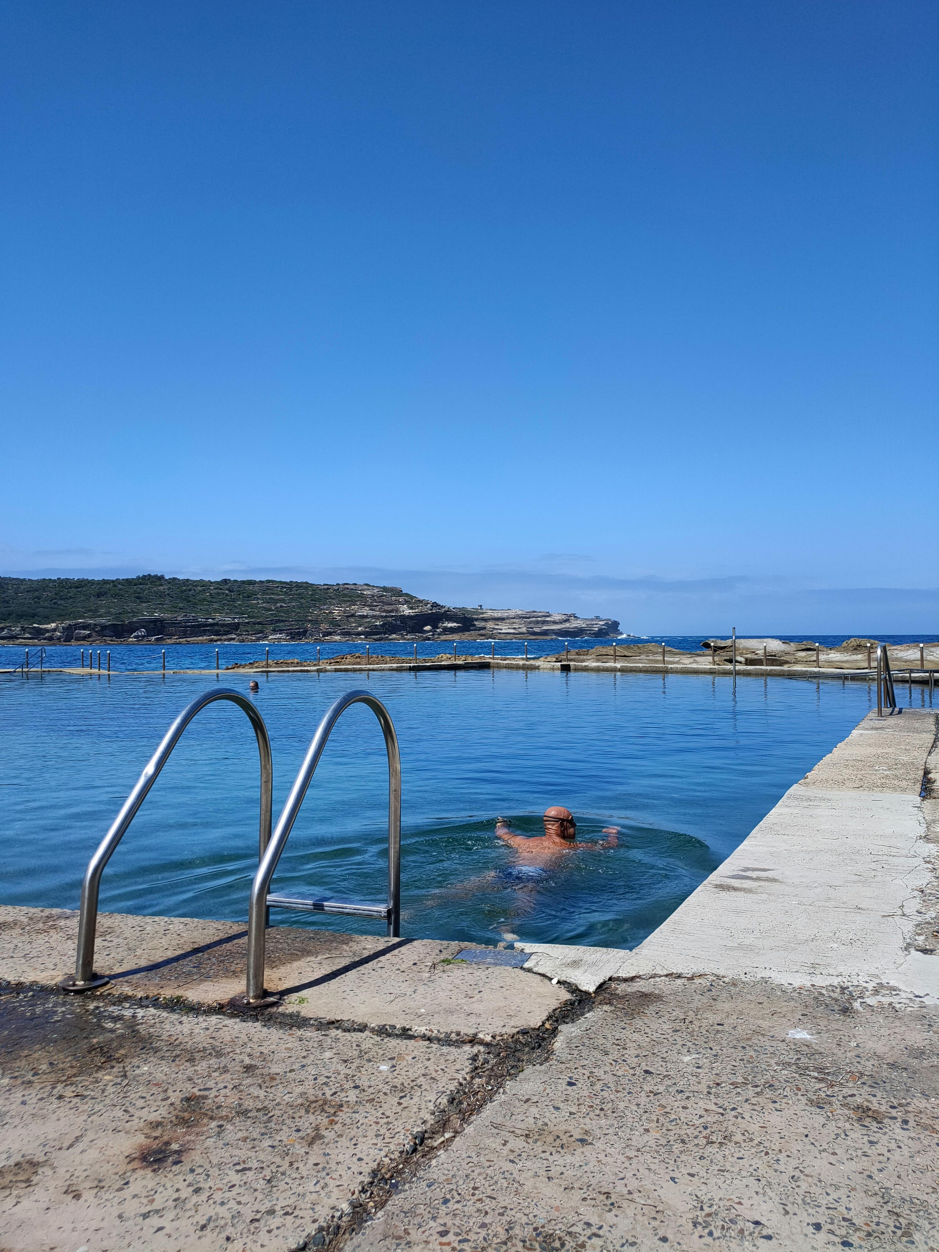 A person enjoys swimming in a coastal ocean pool on a sunny day, overlooking the rocky shoreline.