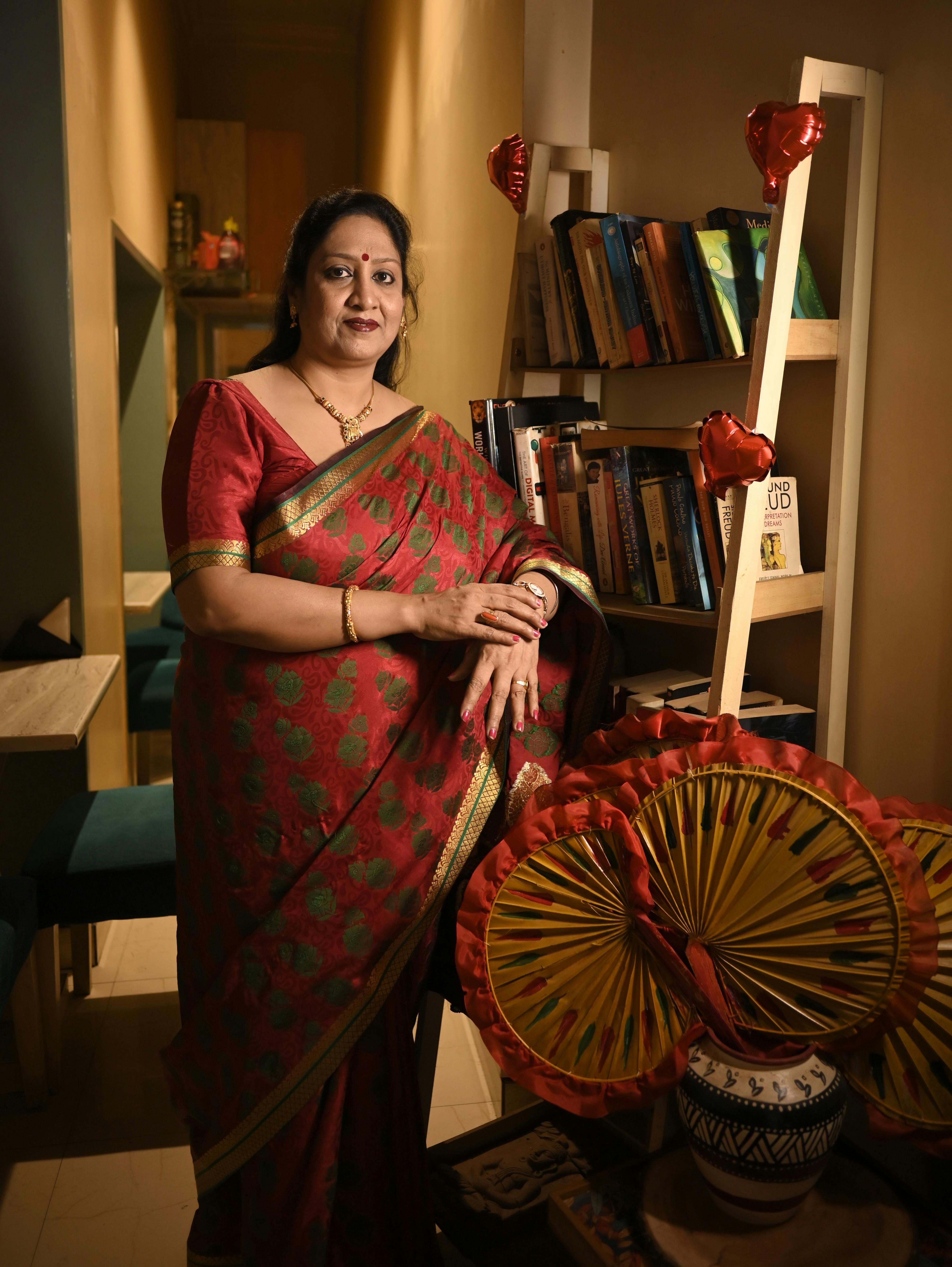 A woman in a red saree posing indoors with decorative items and books around.