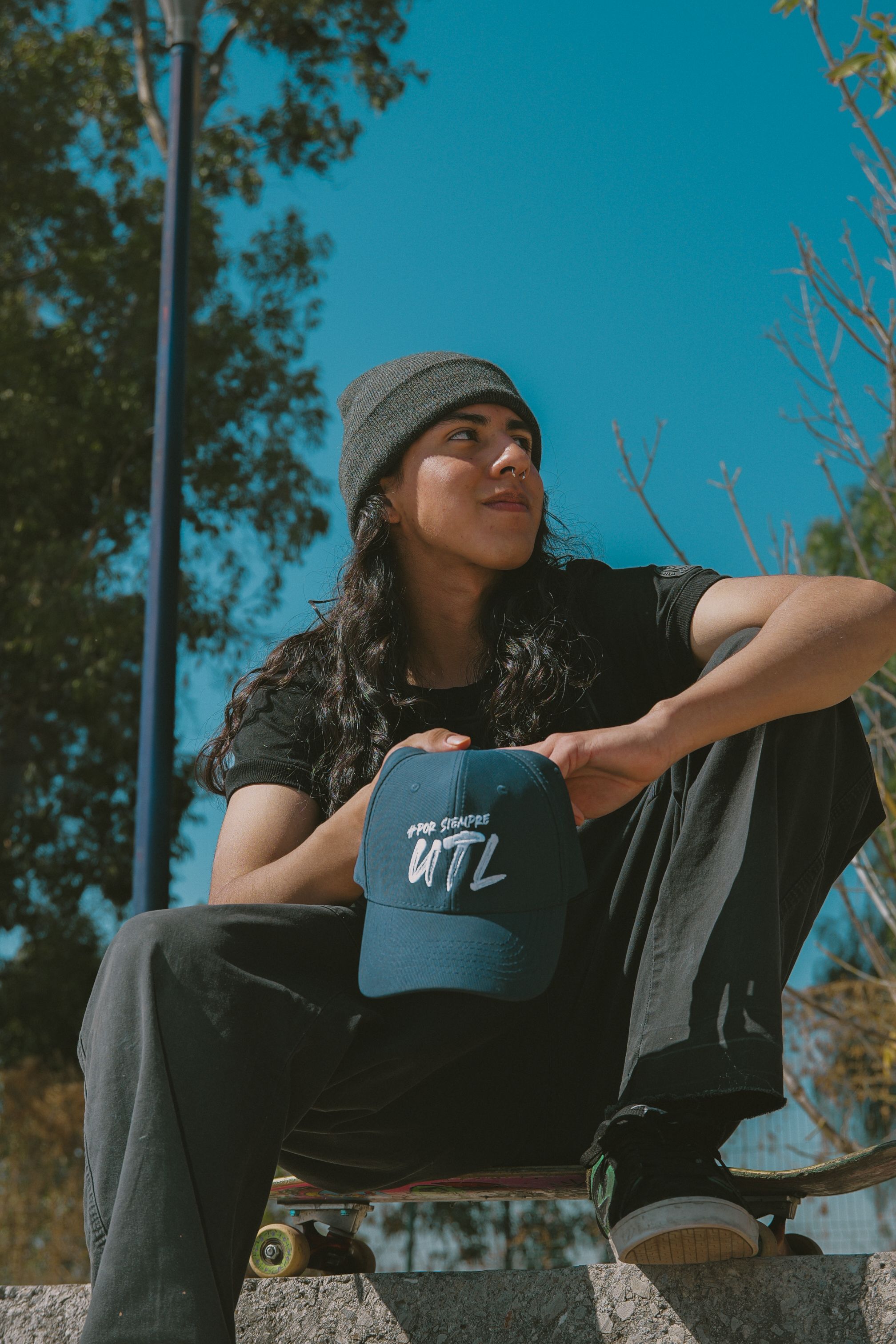 Young skateboarder with long hair and hat sitting on skateboard outdoors.