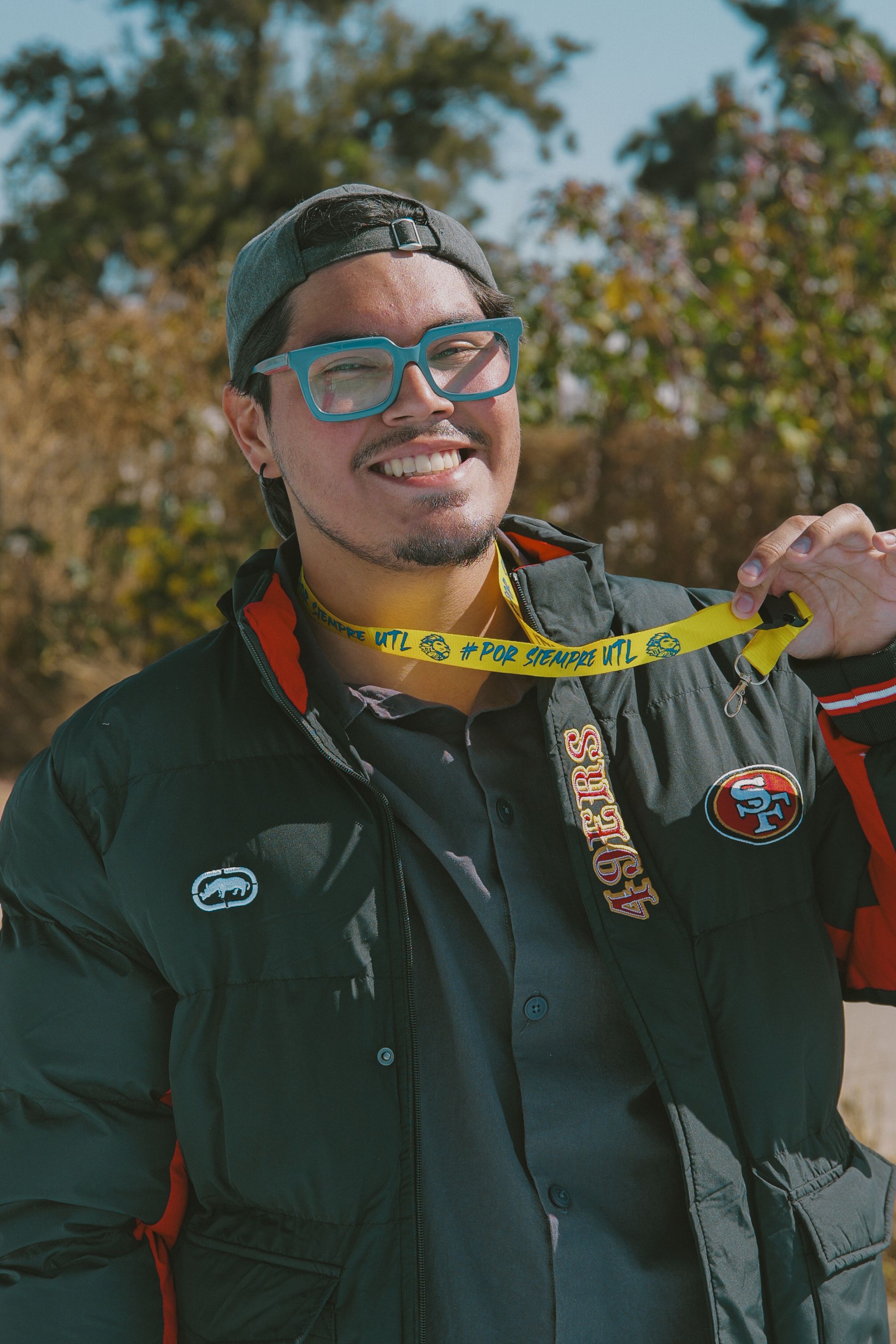 A cheerful man wearing a jacket and cap holds a yellow lanyard while smiling outdoors.