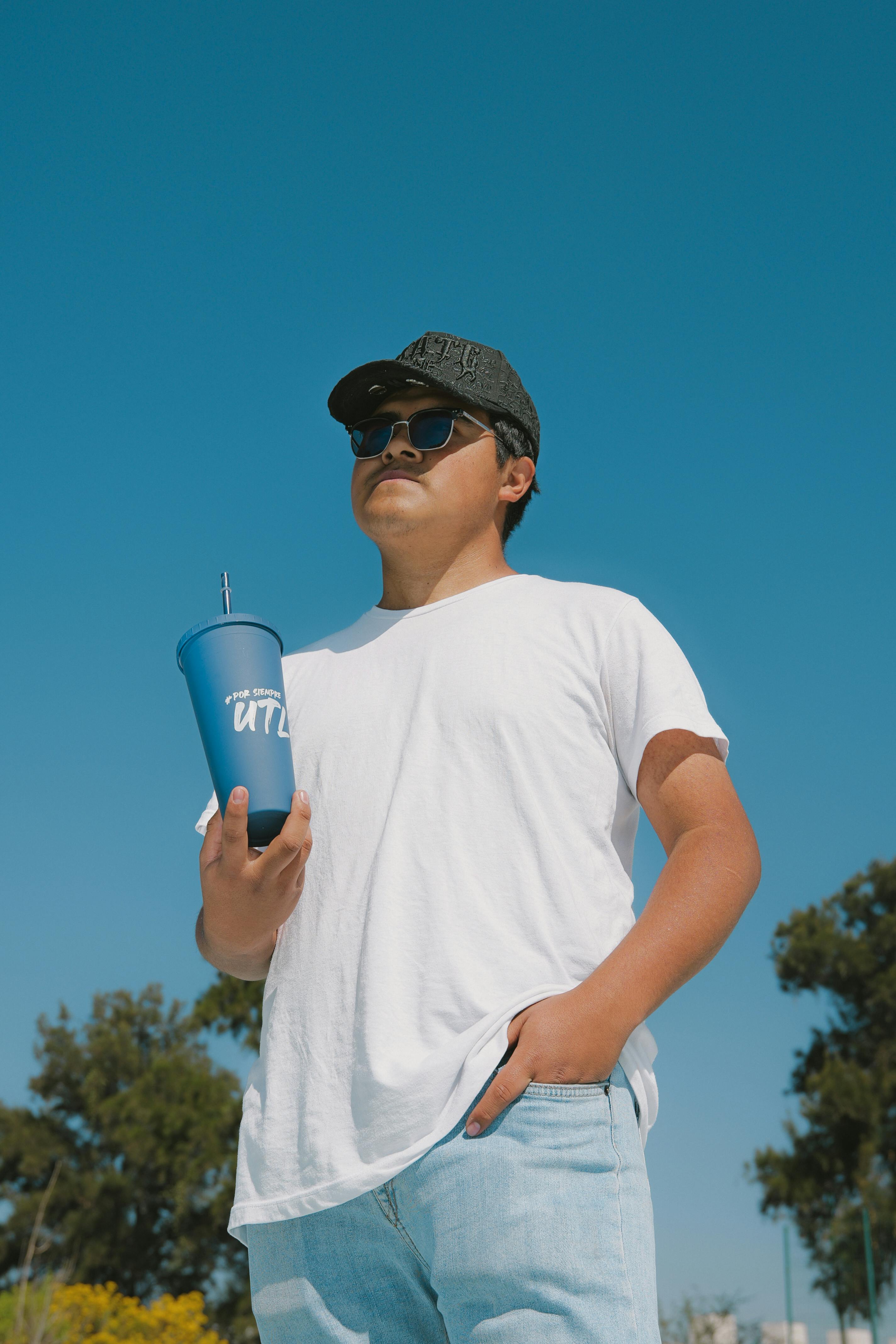 Free A young man in a white shirt and sunglasses holds a blue cup outdoors in León, Guanajuato, Mexico. Stock Photo