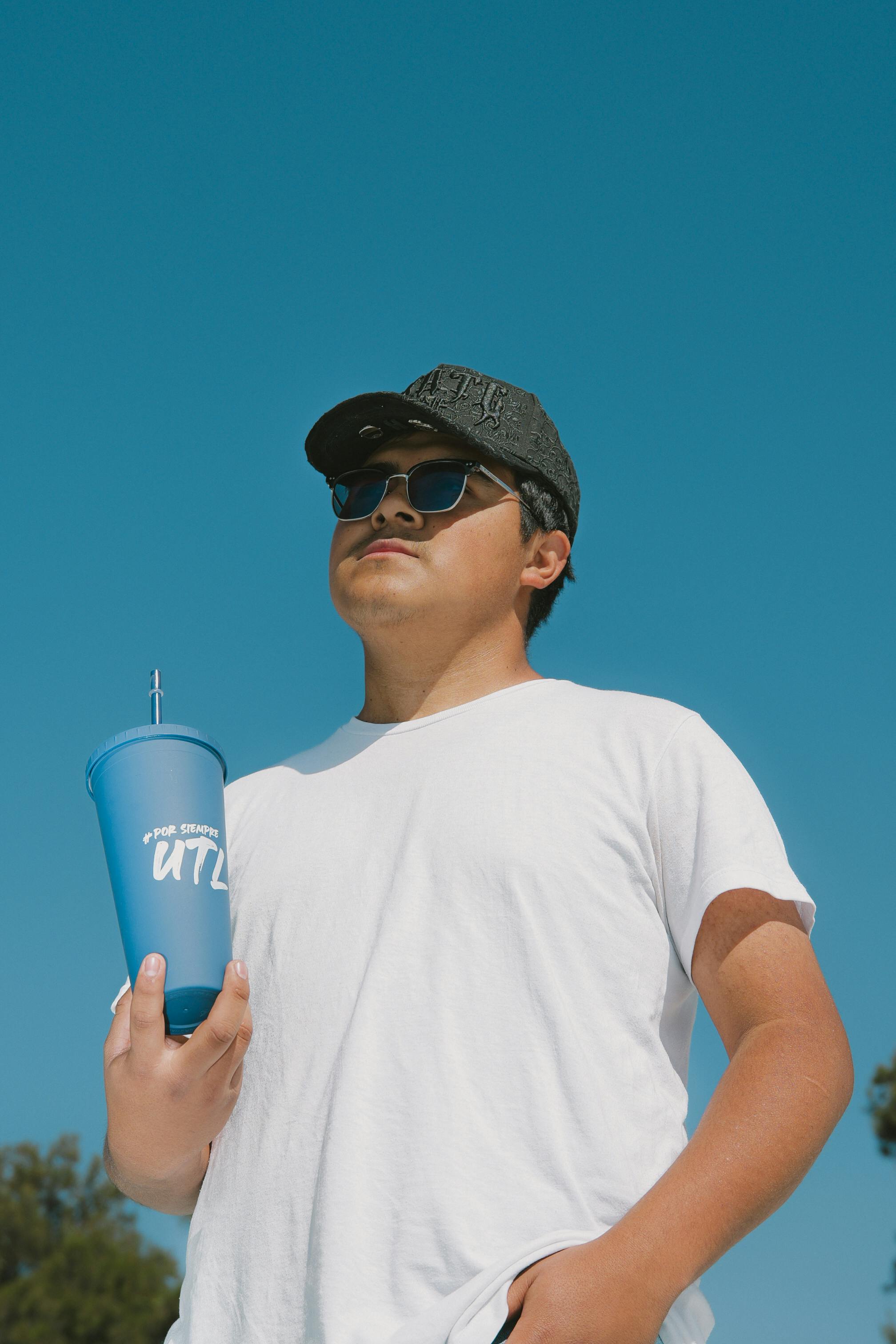 grátis Homem usando boné e óculos escuros segurando uma xícara azul ao ar livre sob um céu azul claro. Foto profissional