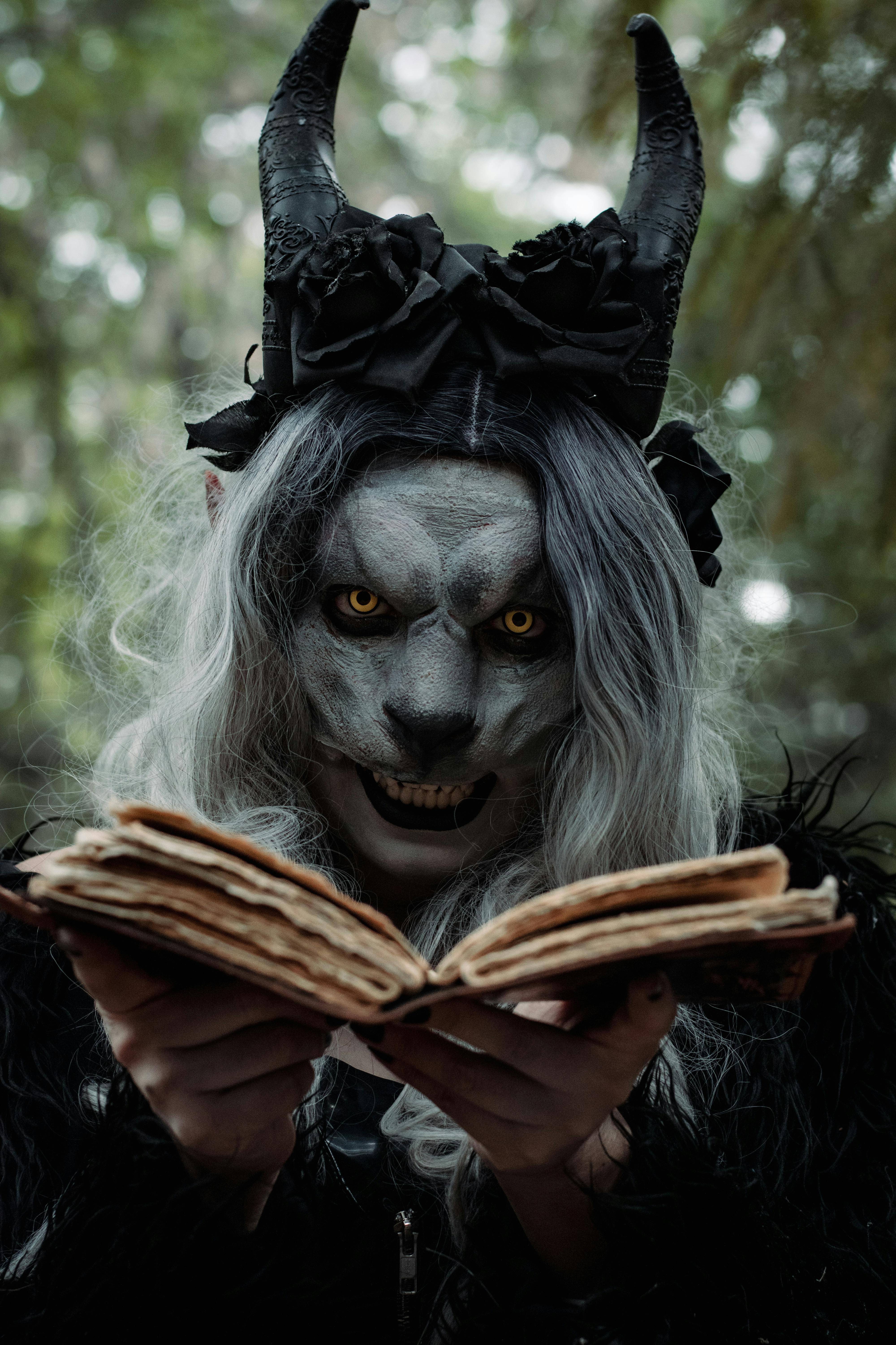 A person in a demonic costume with horns reads an ancient book in a shadowy forest setting.