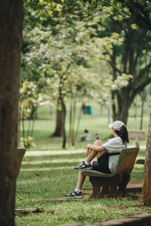 Free A woman sitting leisurely on a bench in a lush green park, enjoying a sunny day. Stock Photo