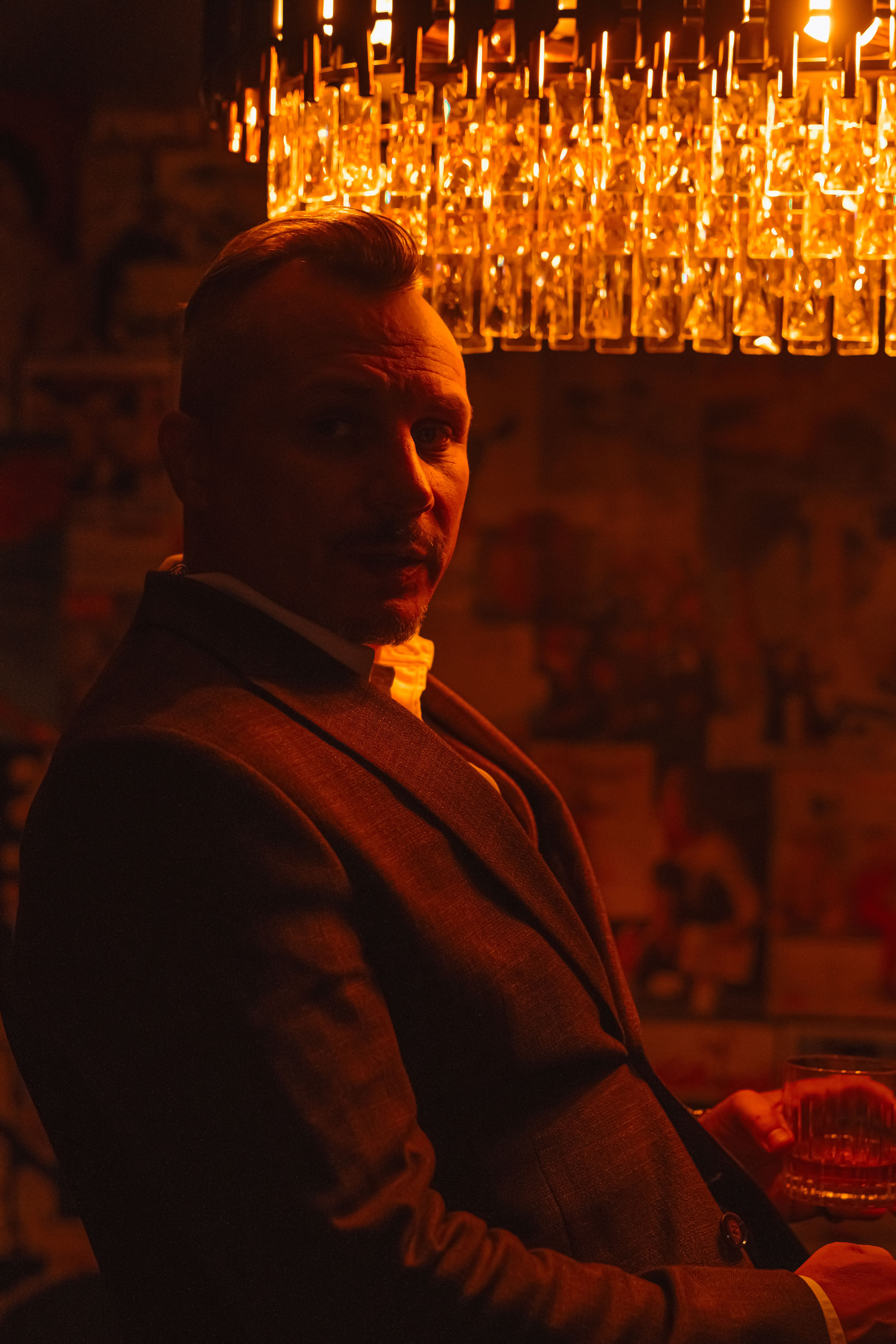Sophisticated man in a dimly lit room, holding a drink under a sparkling chandelier.