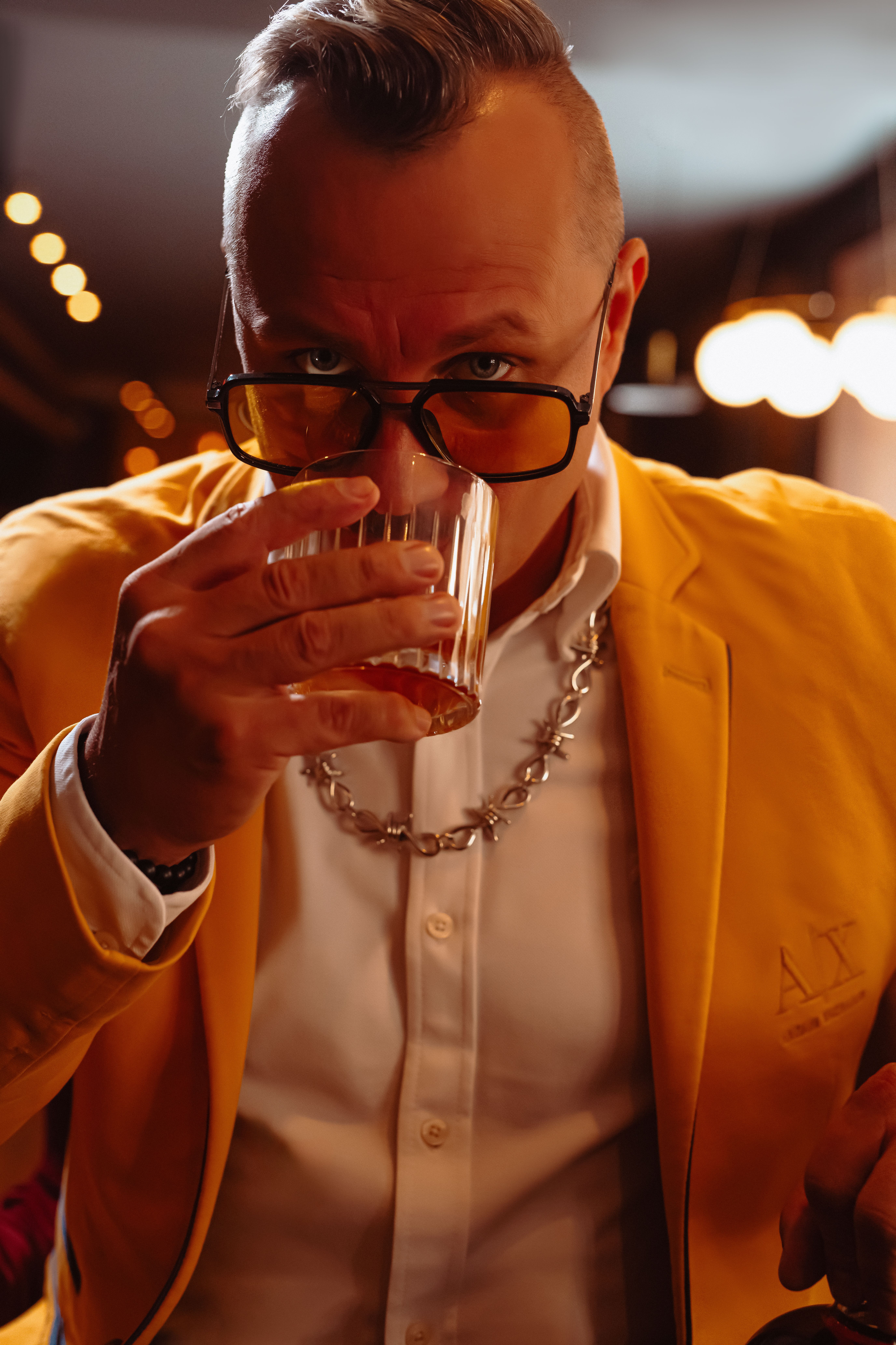 Fashionable man in a yellow blazer sipping a drink at a stylish indoor setting with warm lighting.
