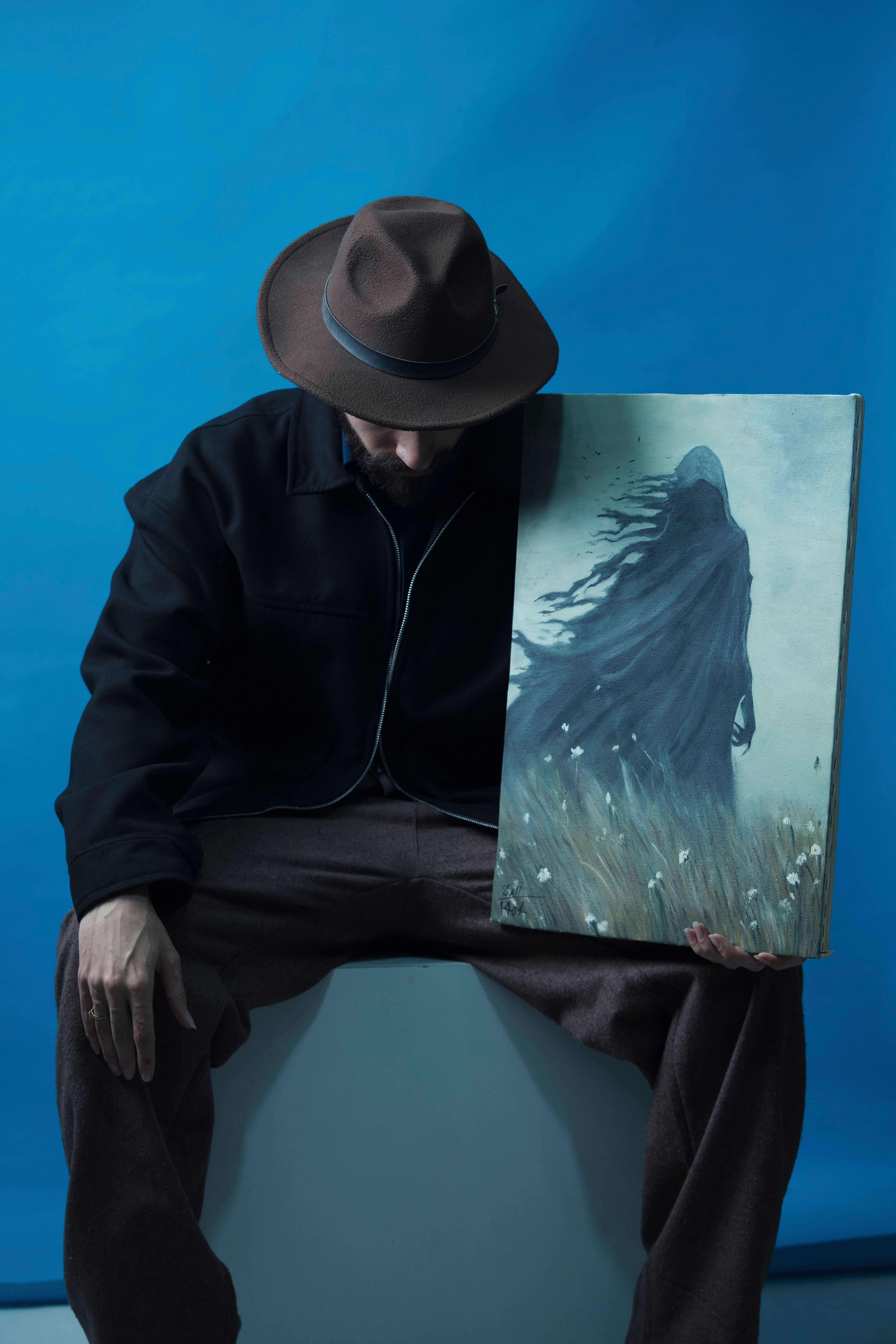 Man in hat holding a mysterious painting with a ghostly figure on a blue background.