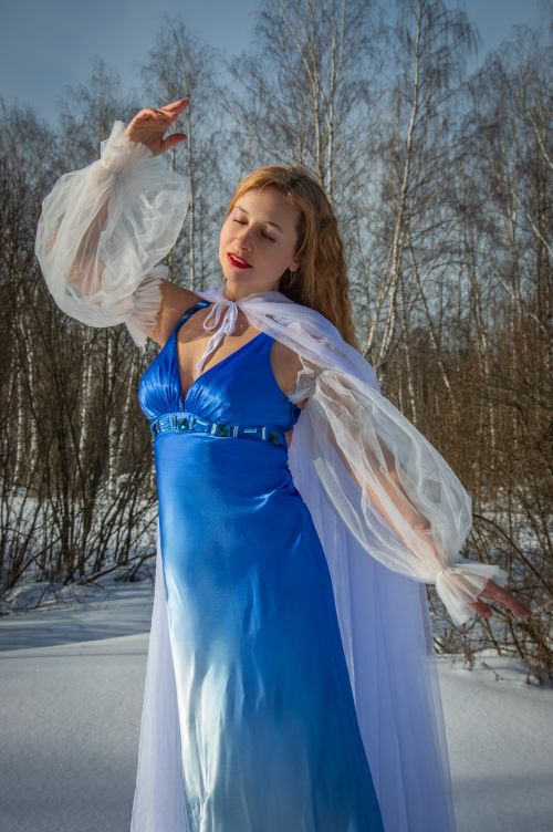 Free Stylish woman in a blue gown poses gracefully against a snowy winter background with bare trees. Stock Photo