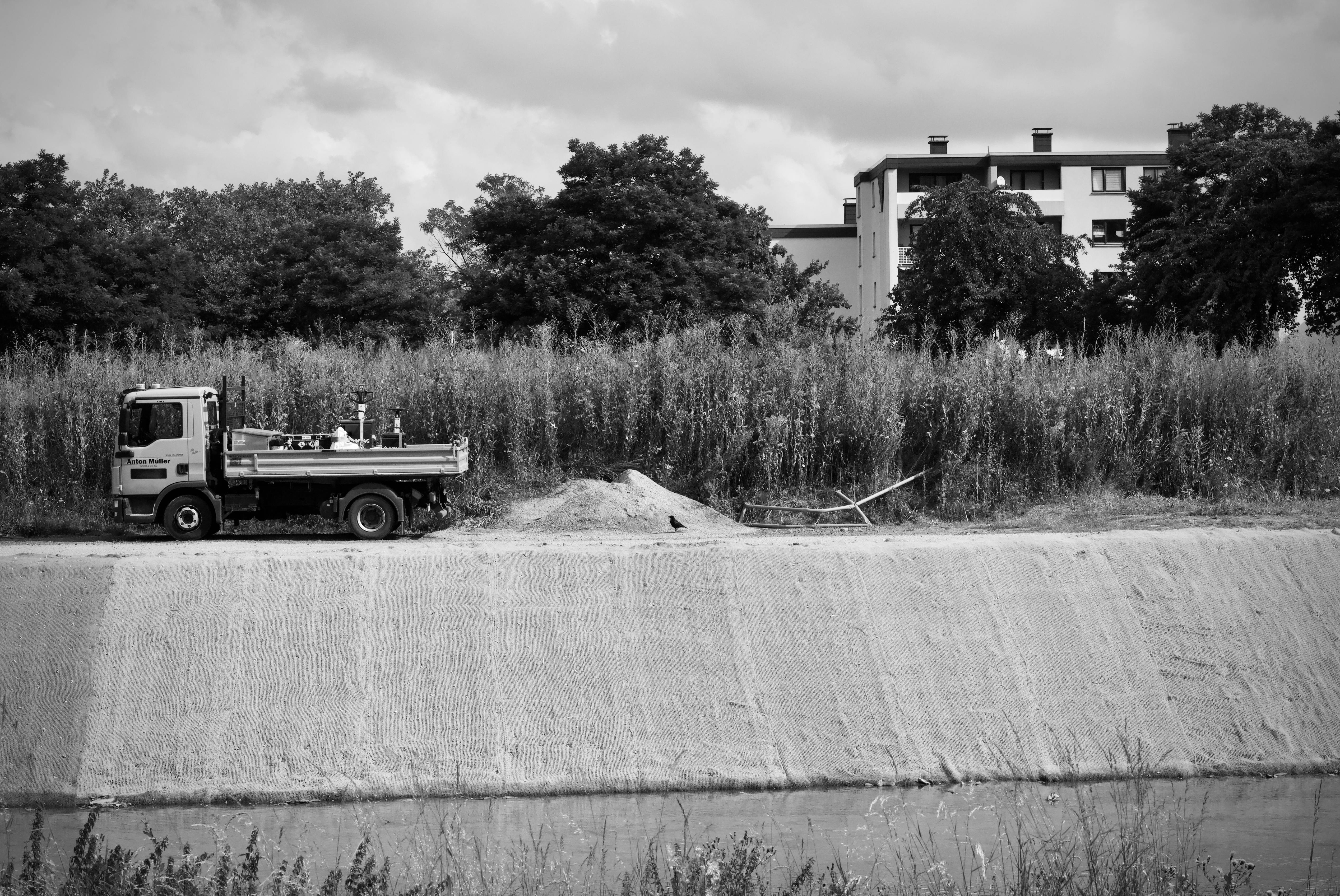 Ücretsiz Ağaçların ve binaların yakınındaki bir inşaat alanında bulunan bir kamyonun siyah beyaz fotoğrafı. Stok Fotoğraflar