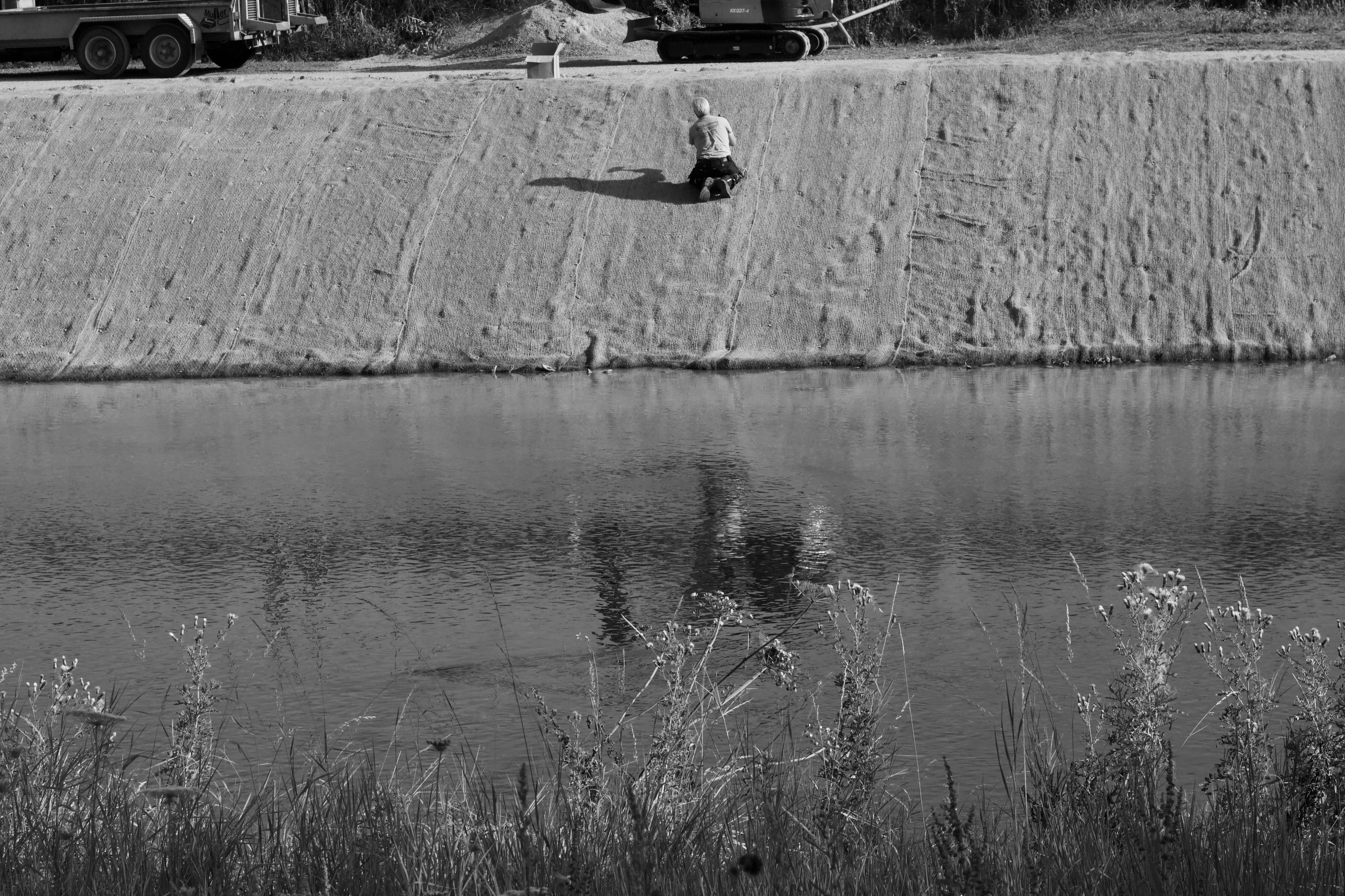 De franc Paisatge monocrom amb un treballador solitari vora una via fluvial, amb maquinària industrial al fons. Foto d'estoc