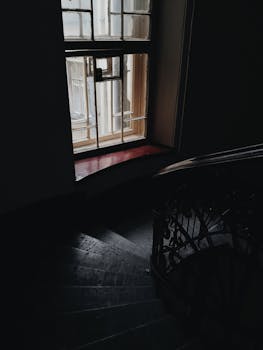 Moody interior shot of a spiral staircase with a window casting soft light, creating a dramatic shadow effect.