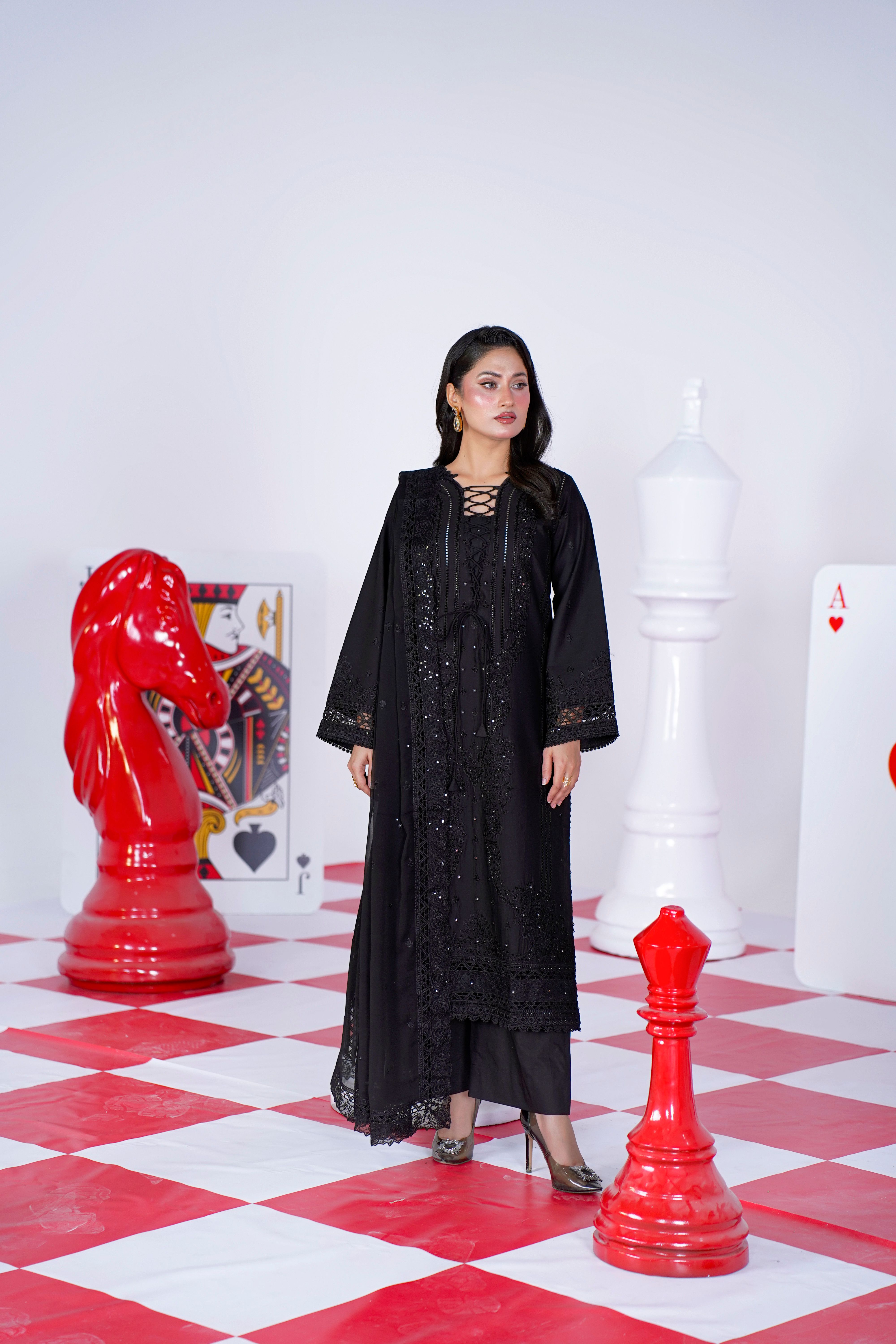 Model in intricate black dress posing with oversized chess pieces indoors.