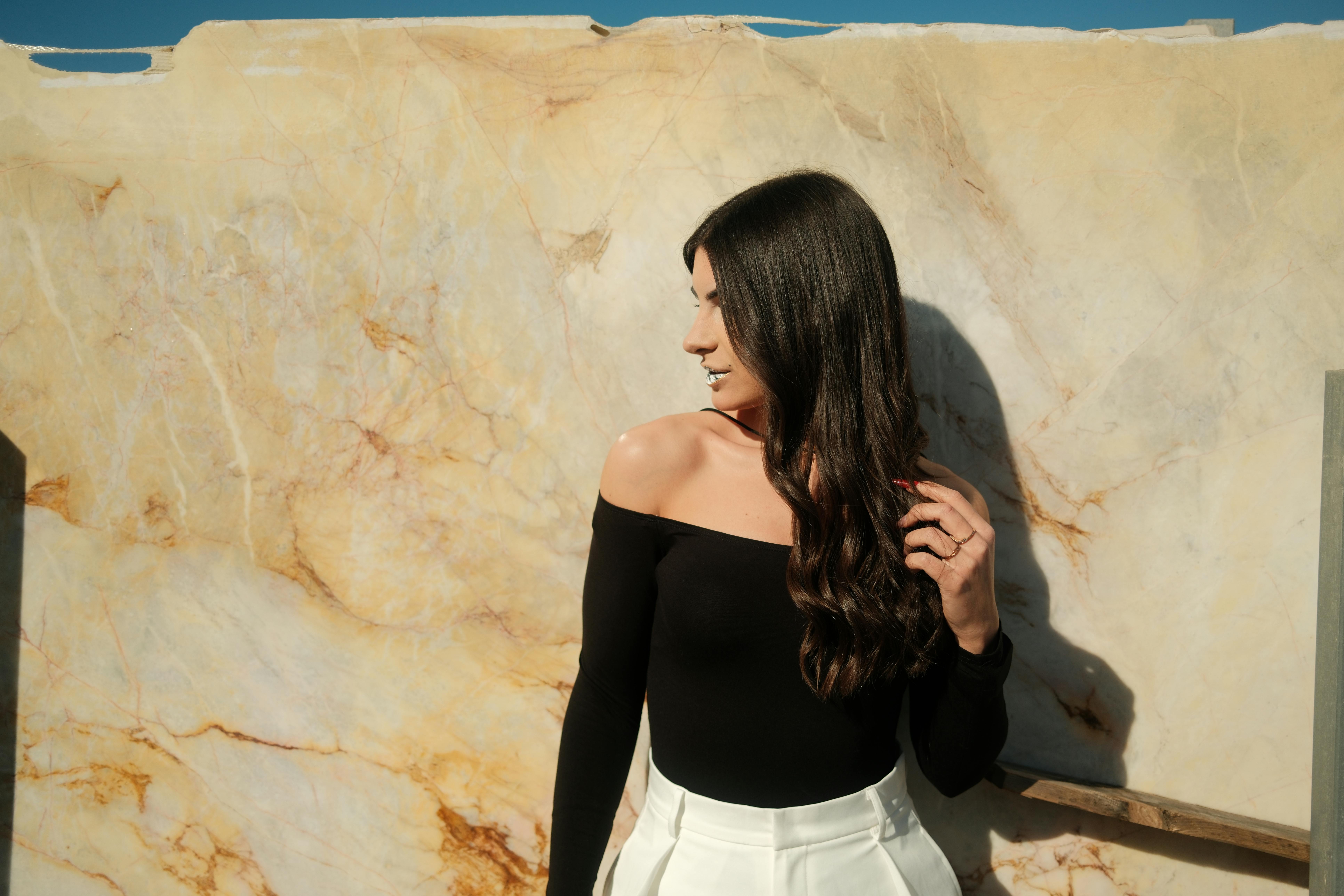Stylish woman in black top and white pants poses against marble wall under bright daylight.