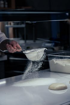 A chef carefully sifting flour onto a kitchen work surface, preparing dough.