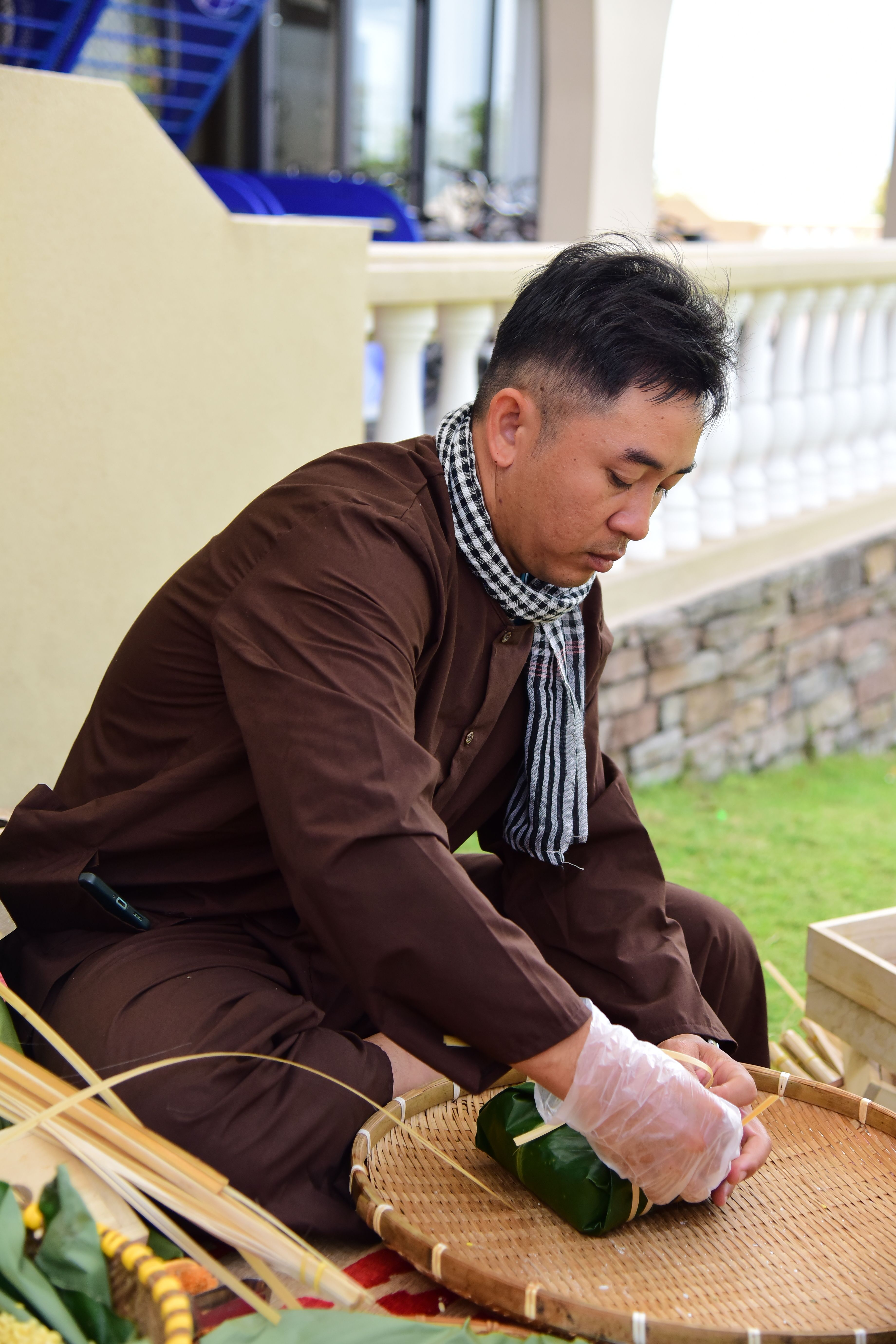 A man in brown attire wraps food in leaves, seated outdoors. Traditional culinary practice.