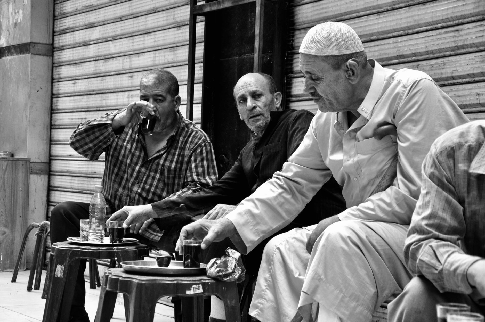 A candid black and white photo of three middle-aged men enjoying tea in an outdoor market setting.