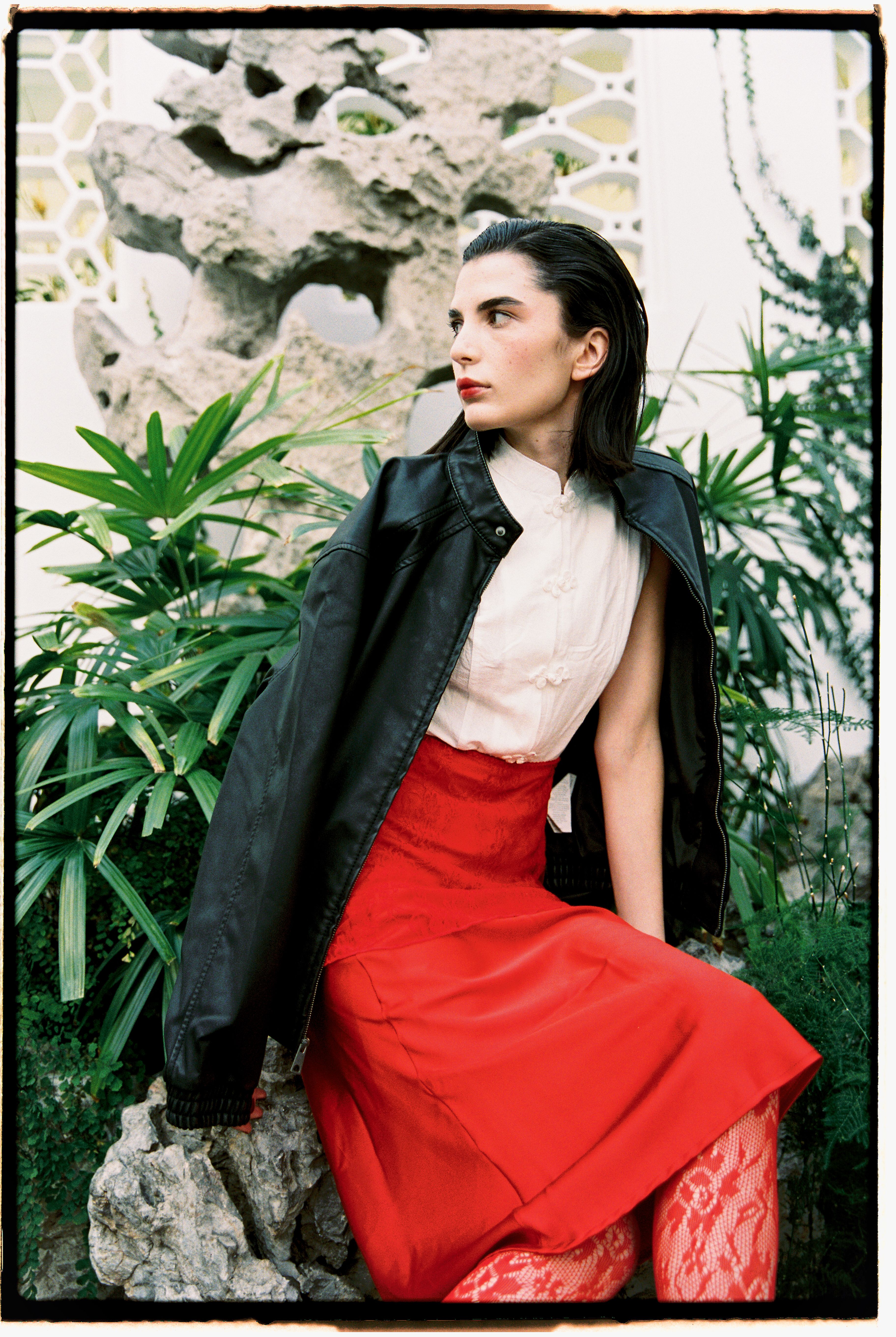 Stylish woman wearing a red skirt and black jacket sitting outdoors in a lush garden.