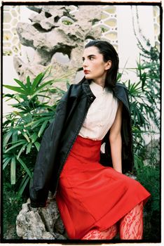 Stylish woman wearing a red skirt and black jacket sitting outdoors in a lush garden.