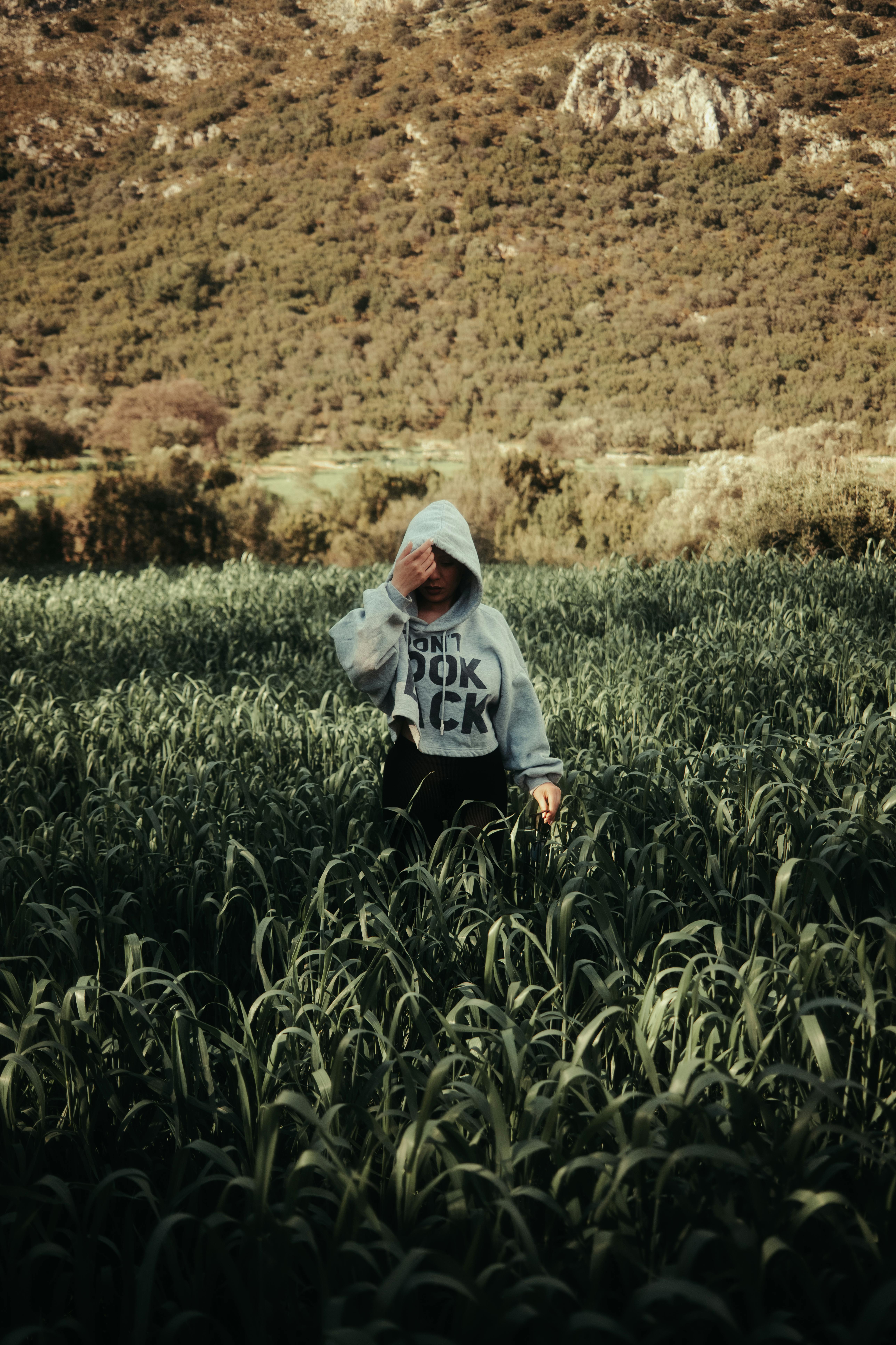 Individual in a hoodie stands amidst tall plants with hills in the background, evoking solitude and nature.