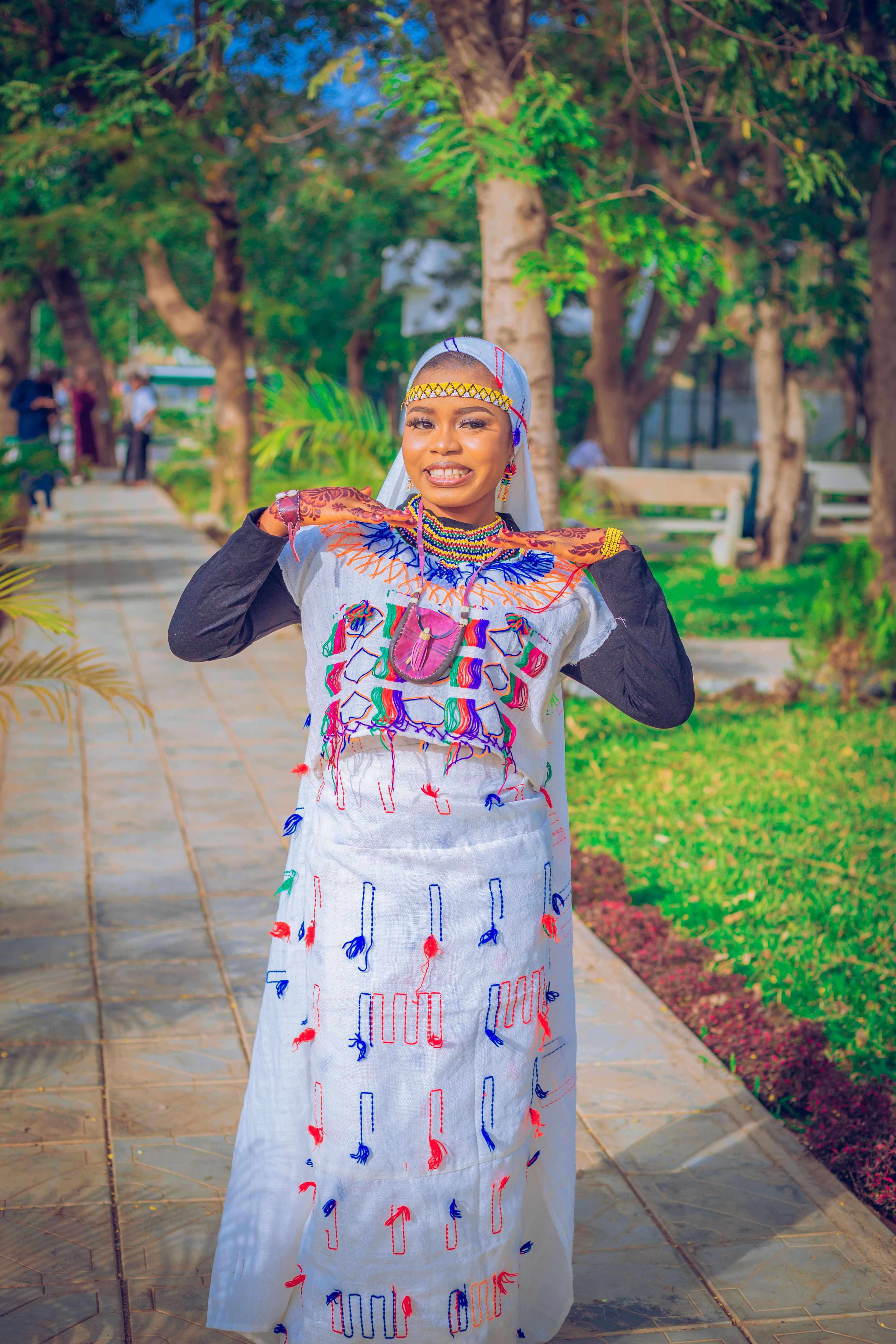 Smiling woman in vibrant traditional African attire posing in a sunny park setting, symbolizing cultural beauty.