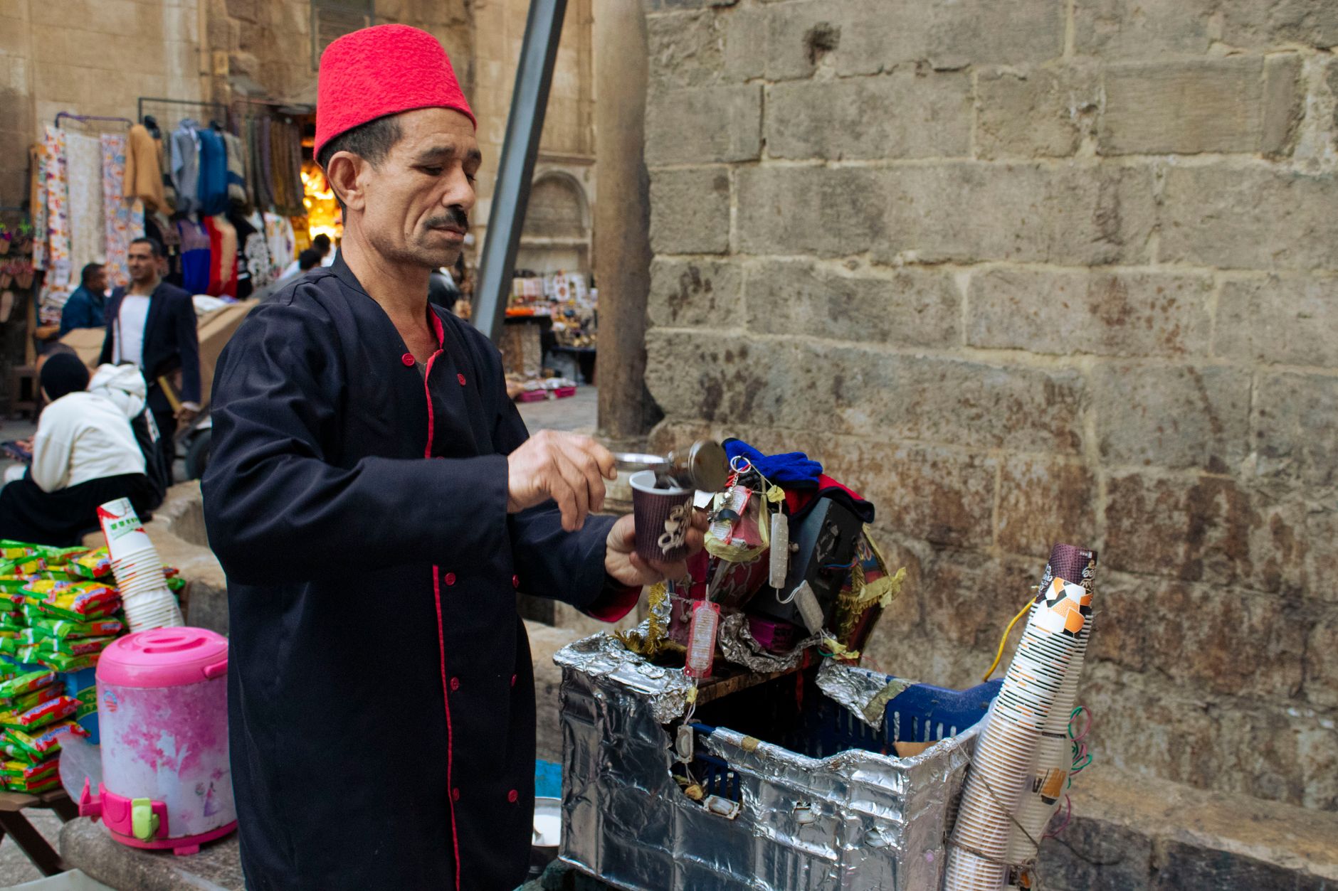 A street vendor in a red fez prepares a traditional drink at a bustling market.