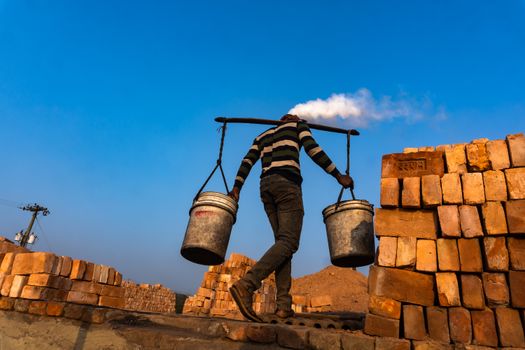 A laborer carries buckets on a yoke through a brick yard under a clear blue sky, showcasing manual labor.