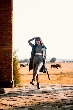 Fashionable woman strolls outdoor in rustic setting, surrounded by grazing cattle in fall ambiance.