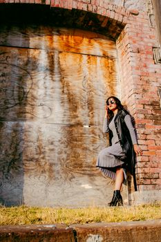 A stylish woman poses against a textured wall in a sunlit outdoor setting, wearing a chic outfit.
