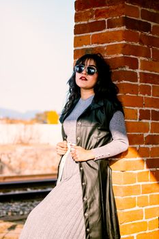 Stylish woman wearing sunglasses and leather vest leaning against a brick wall in sunny outdoors.
