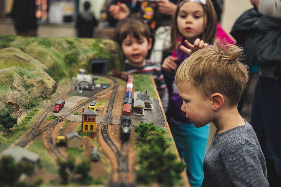 Selective Focus Photo of Boy Standing Near Miniature Train Toys