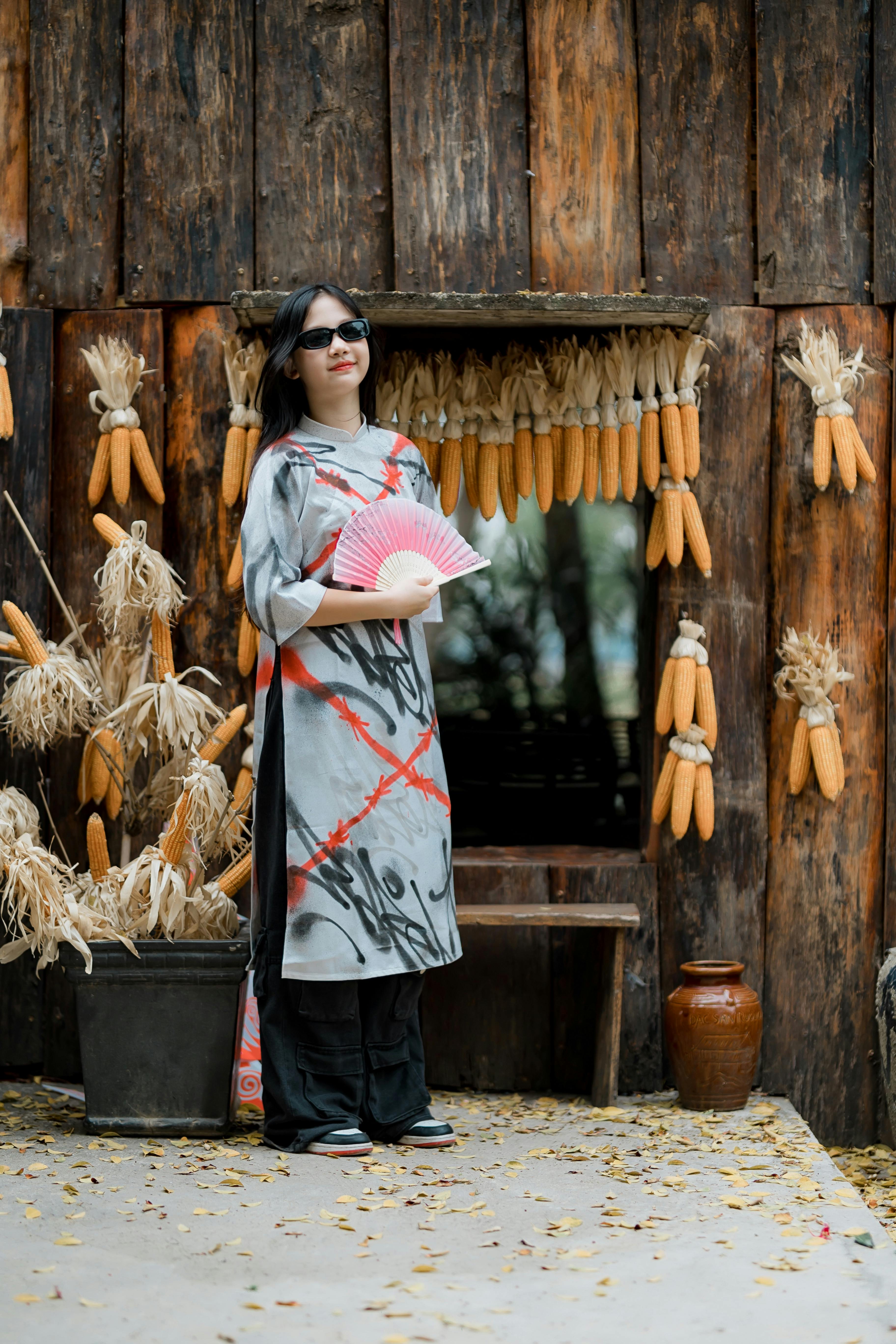 Woman in sunglasses posing with a fan against a rustic corn-decorated wall.
