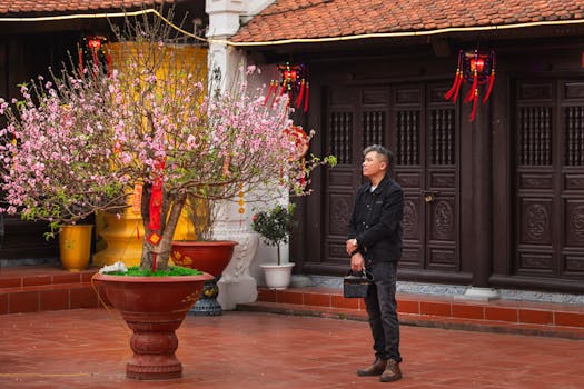 A man stands beside a blossoming peach tree with red decorations in a traditional Asian courtyard.
