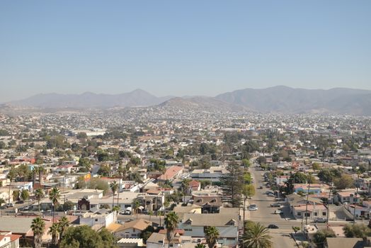 Aerial daytime view of Tijuana's cityscape with mountainous backdrop under a clear sky.