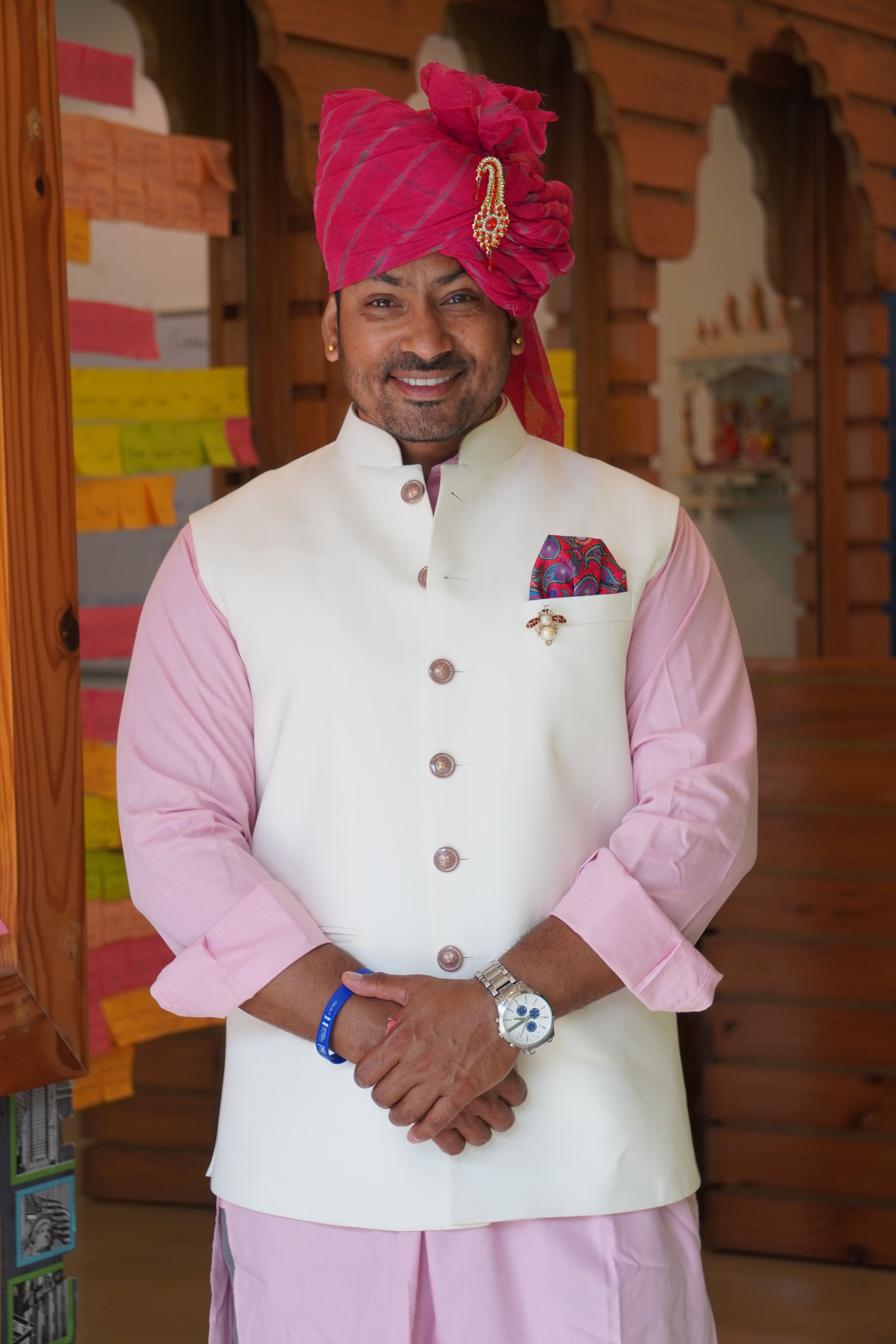 Smiling man in traditional white and pink attire with a pink turban, standing indoors.
