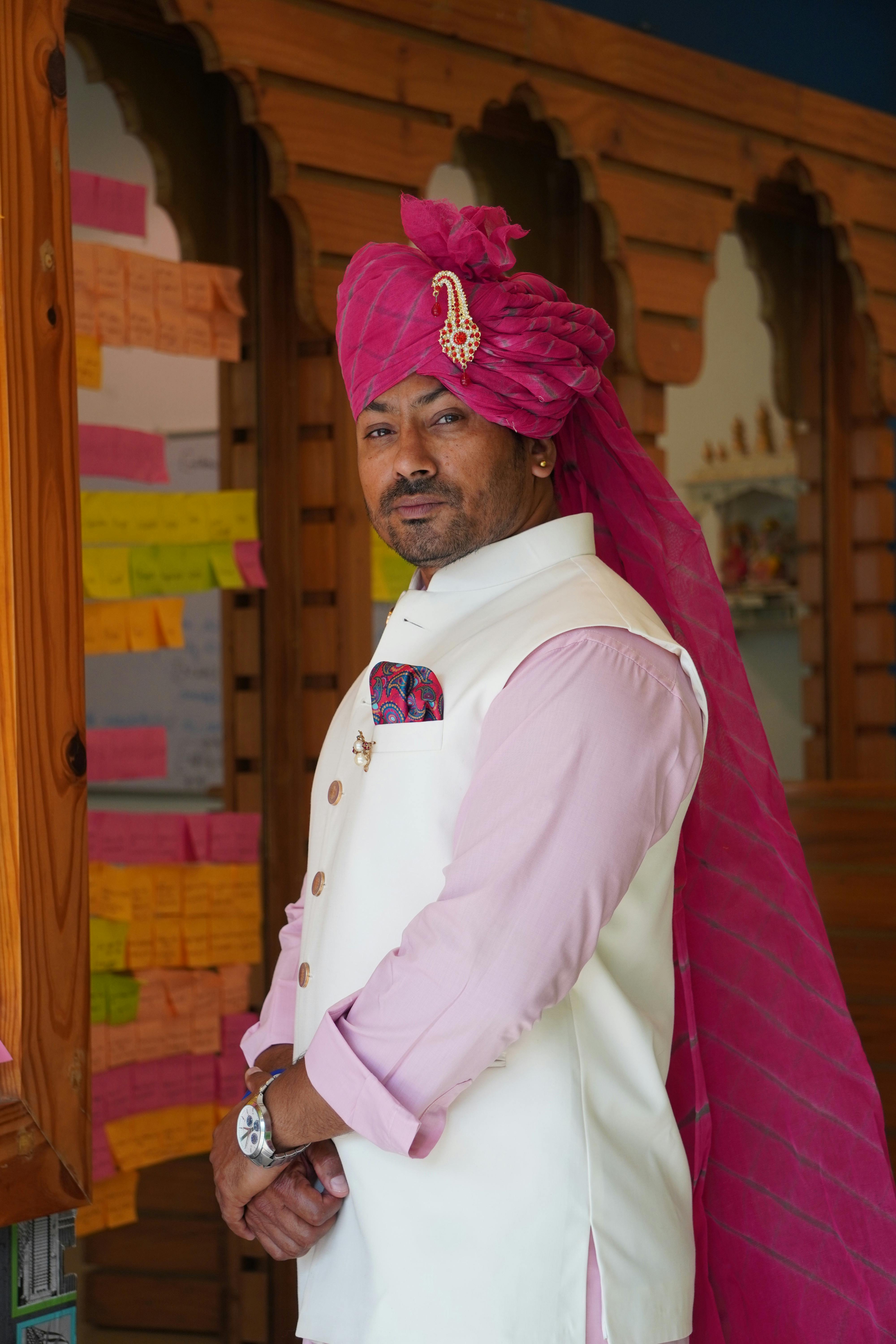 A man in a traditional pink turban and white attire posing indoors, reflecting cultural elegance.