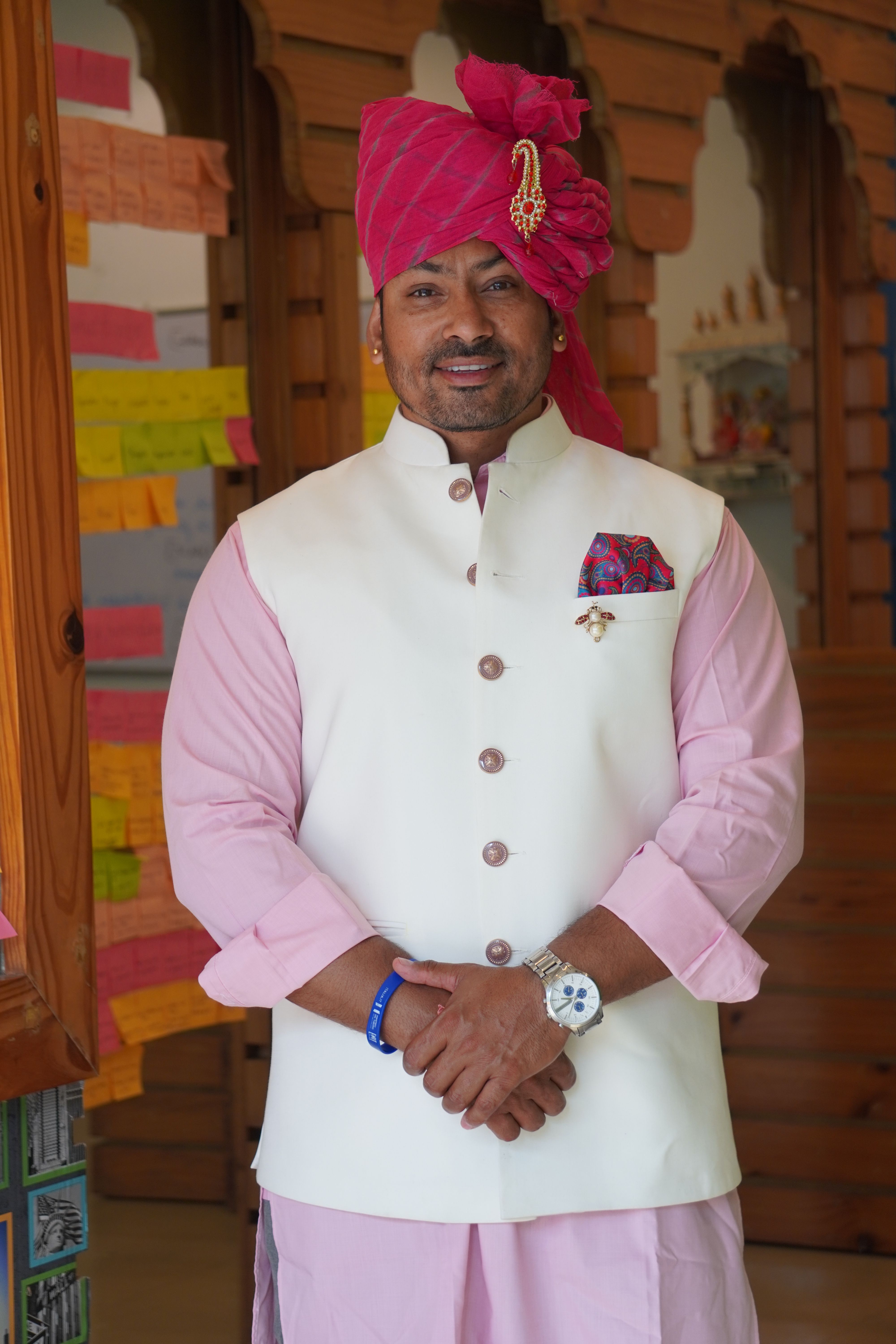 Man wearing traditional Indian clothing and turban standing indoors, smiling warmly.