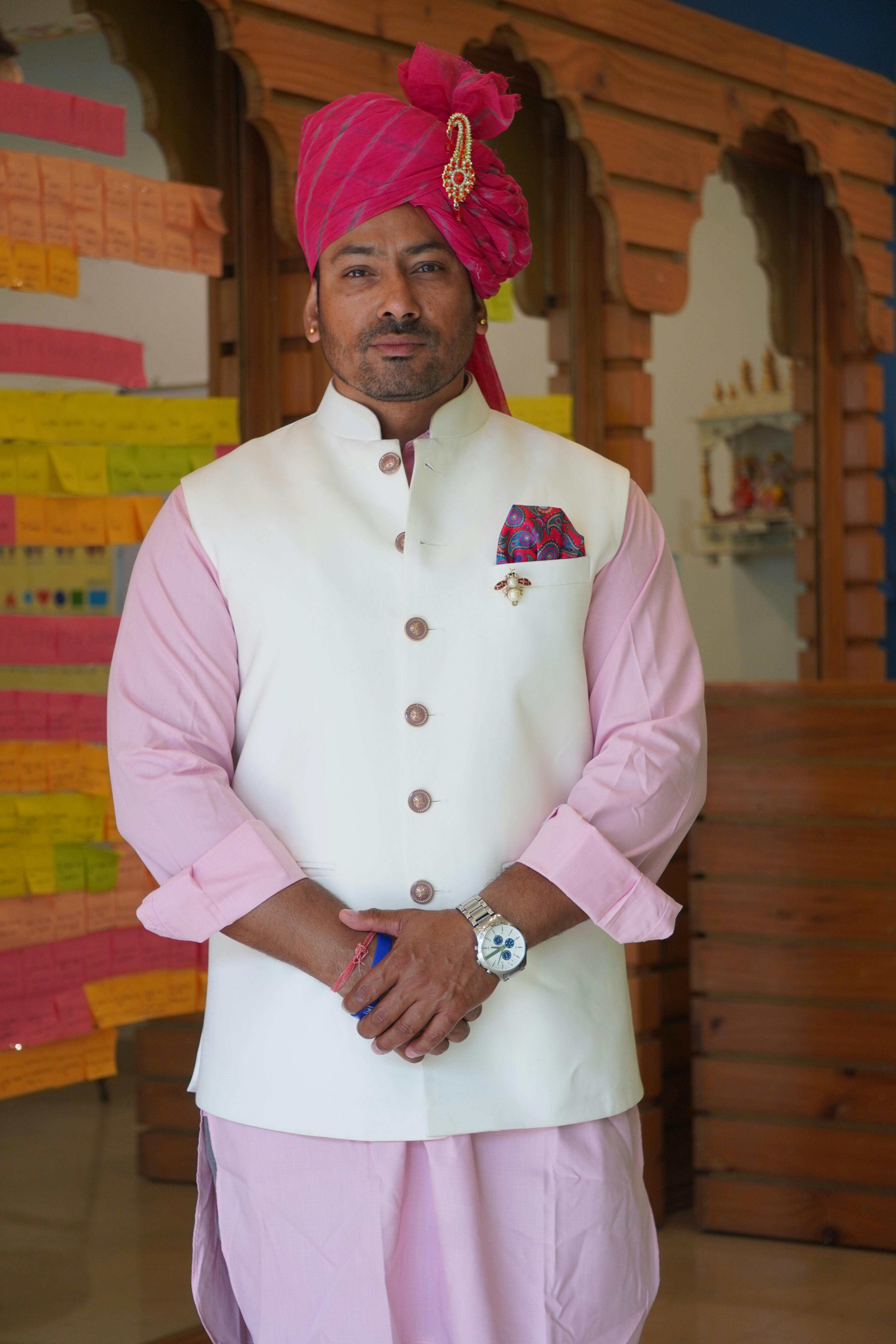 Man in pink and white traditional Indian attire with turban, standing indoors among colorful decor.
