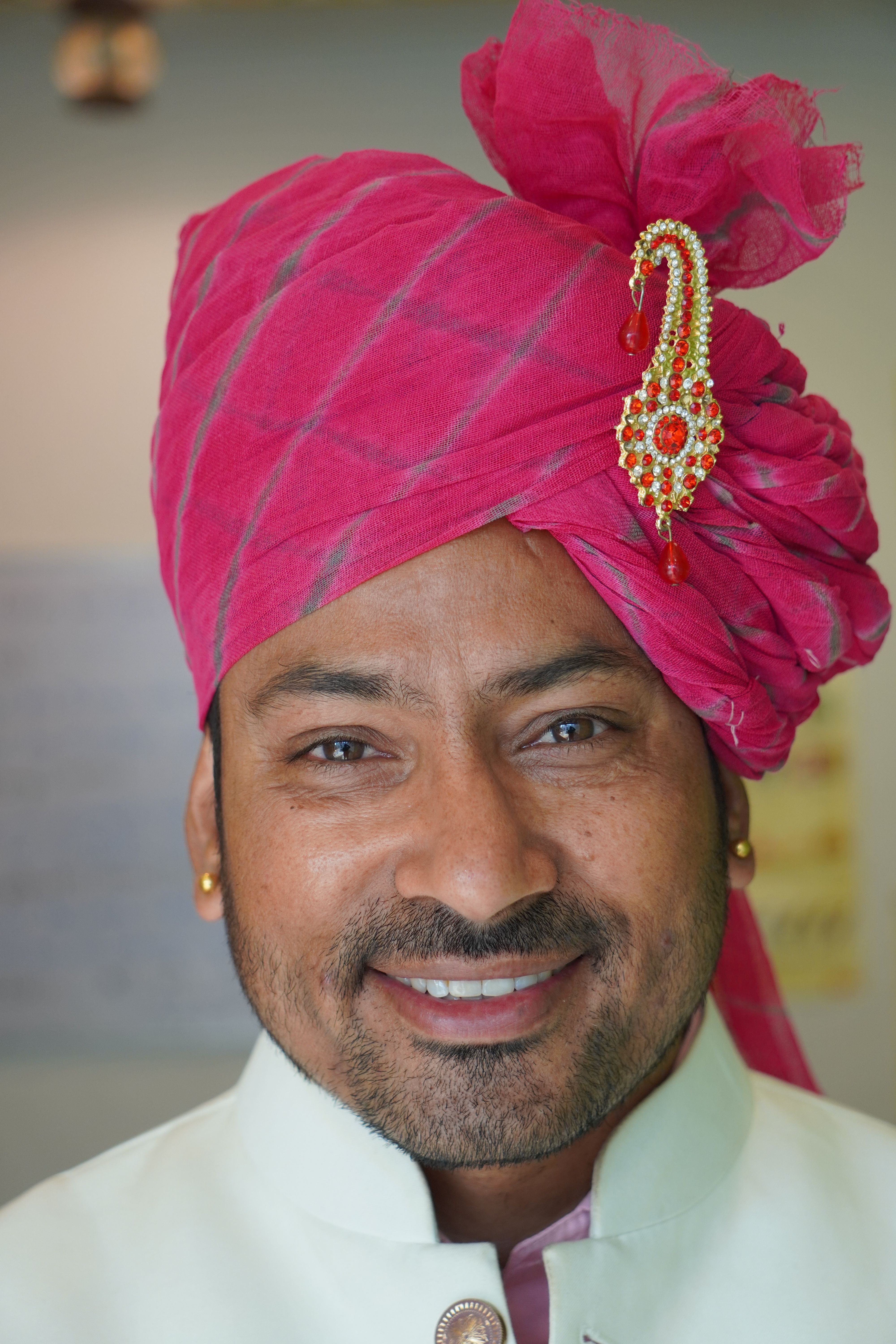 A cheerful man wearing a vibrant pink turban and white traditional attire indoors.