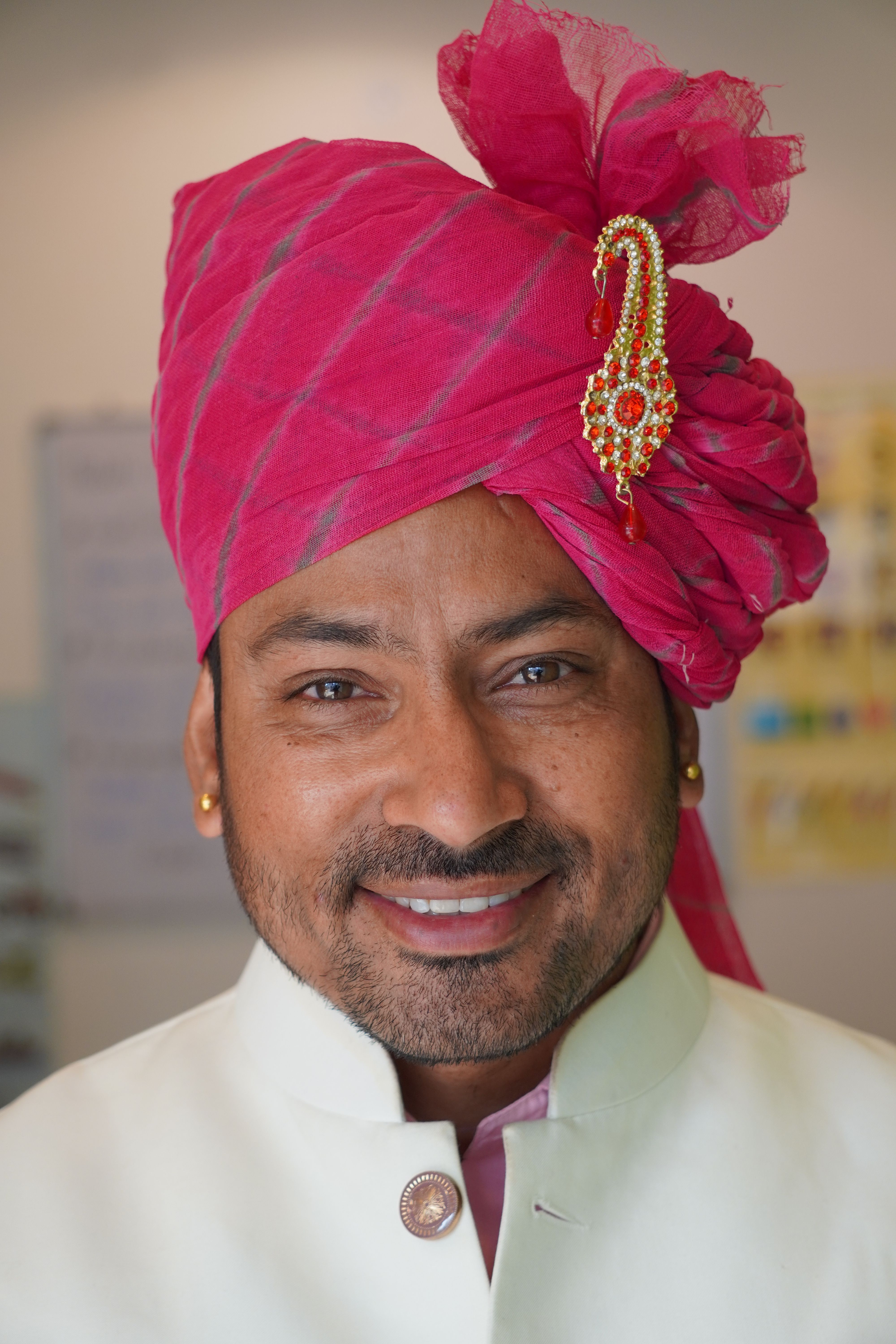 A man wearing a vibrant pink turban and white traditional attire, smiling warmly indoors.