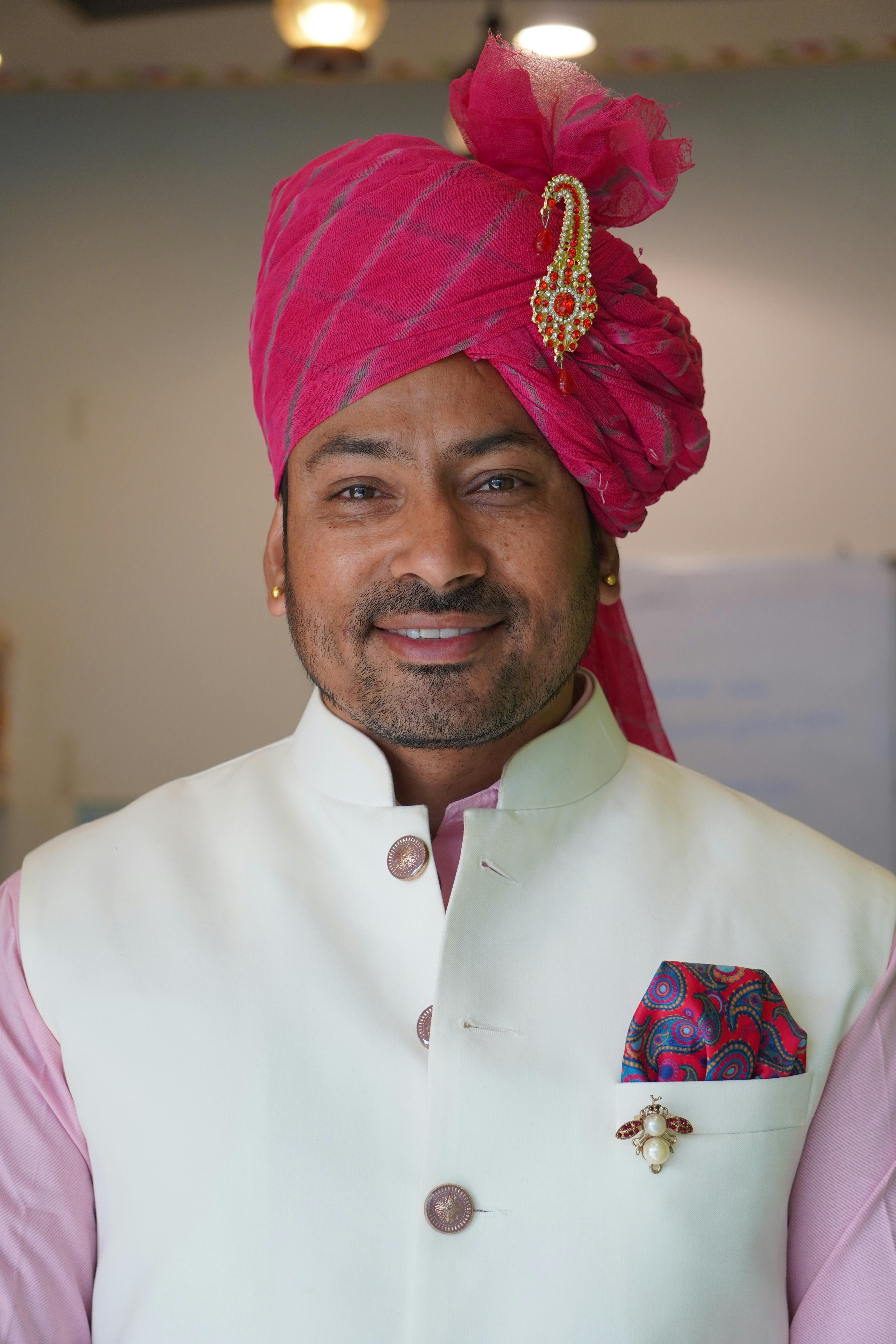 Man smiling in traditional attire with pink turban, indoors setting.